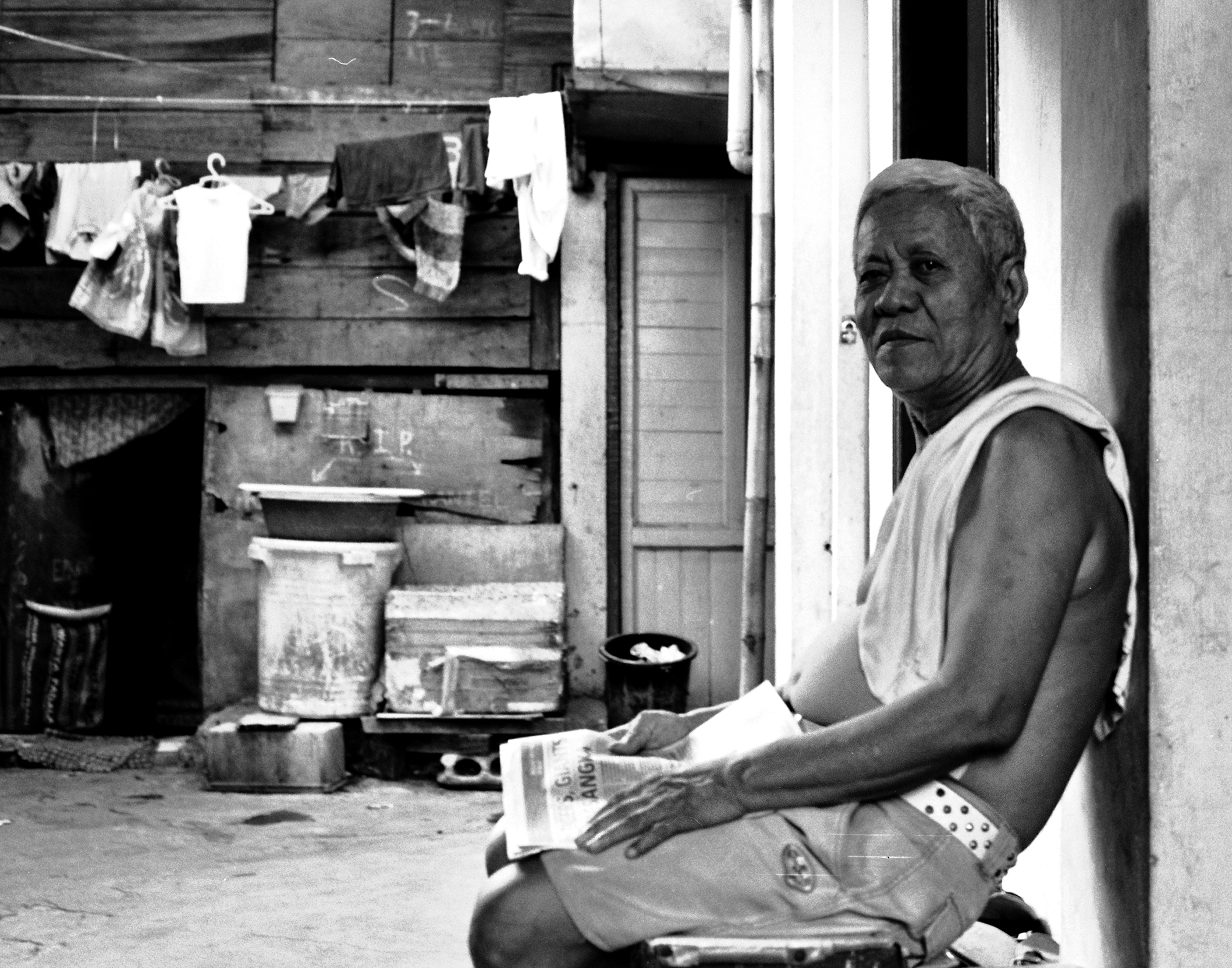 Elderly man in tank top seated reading newspaper with laundry hanging on clothesline behind him in modest dwelling