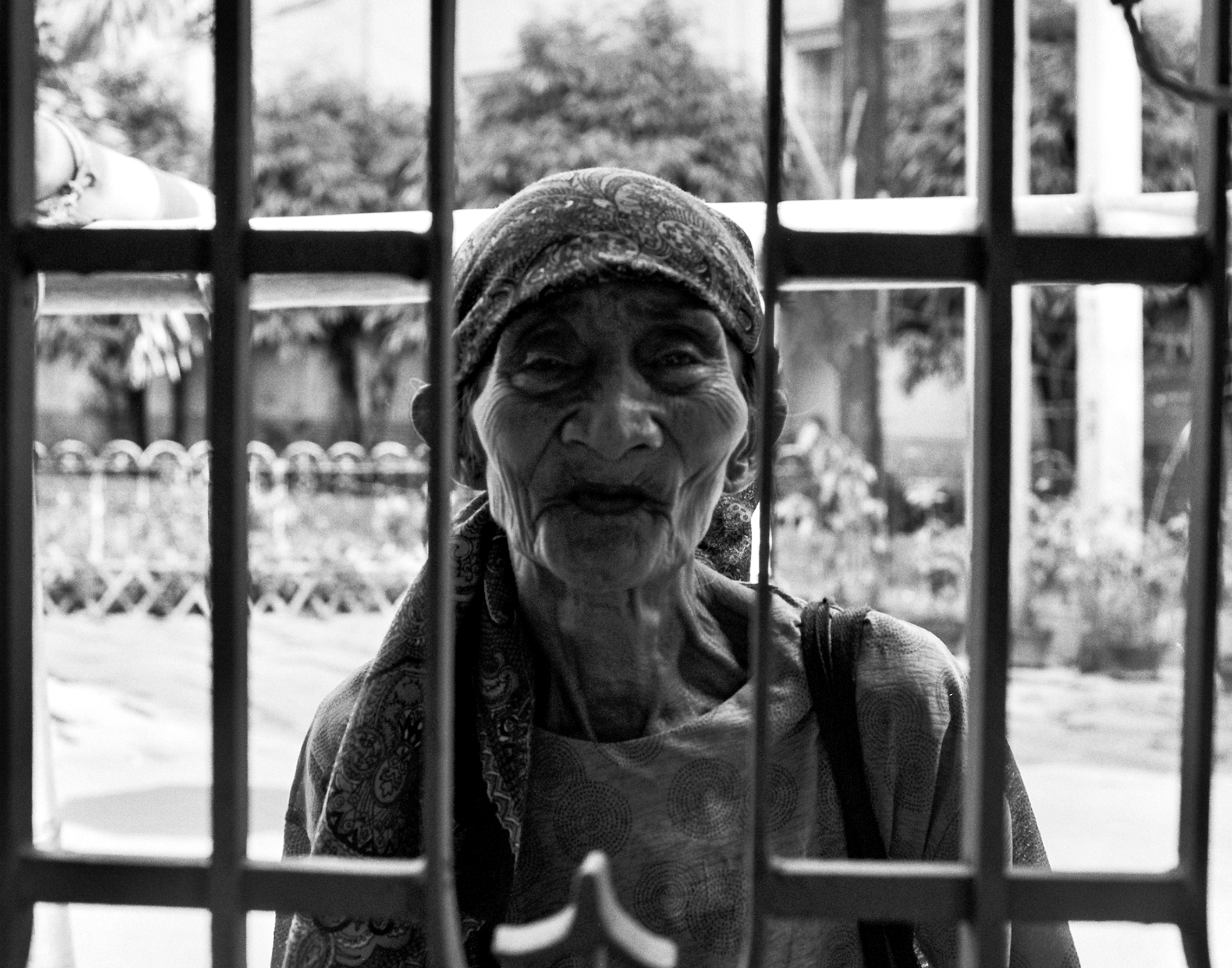 Elderly woman with weathered face and headscarf viewed through metal bars with direct powerful gaze