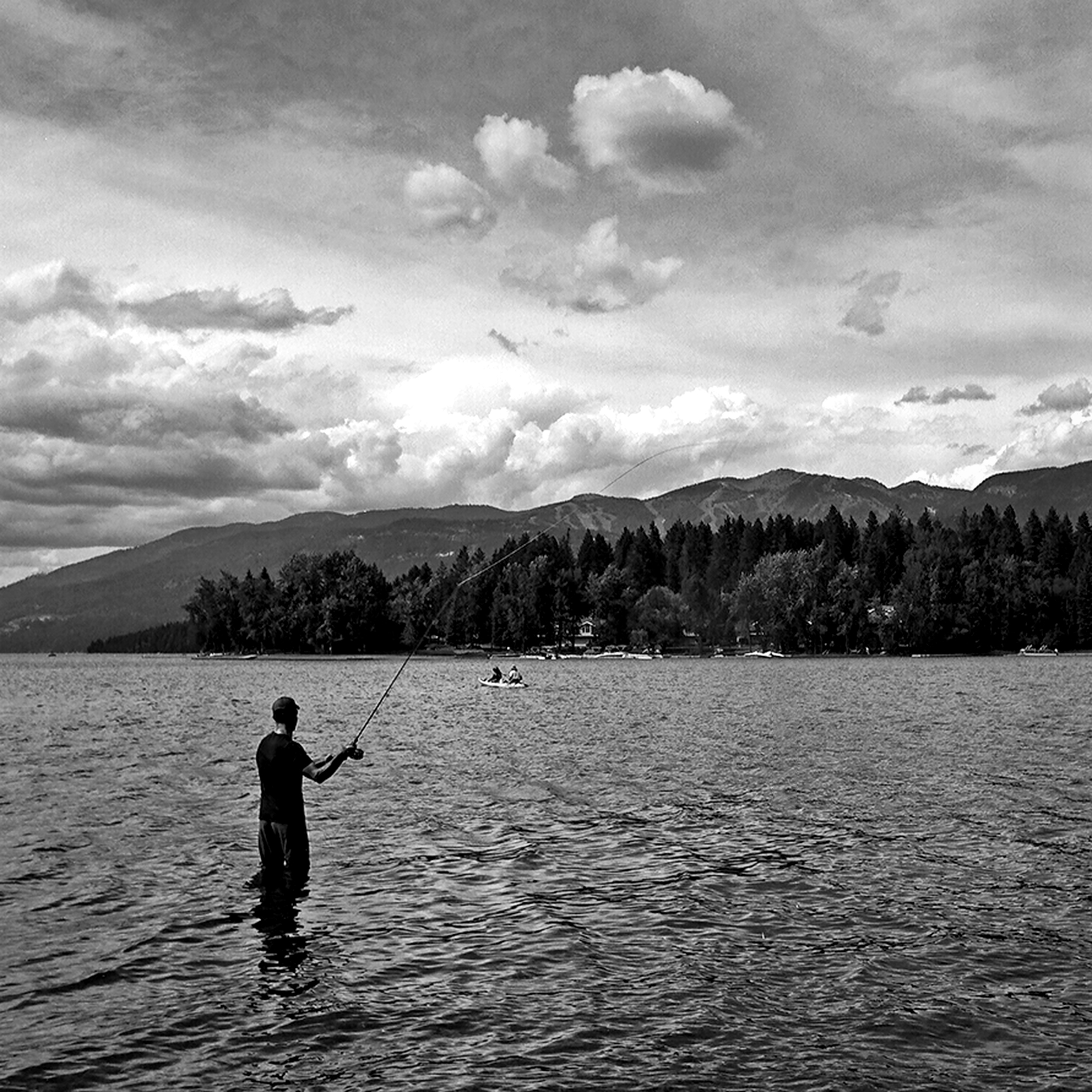 Fly fisherman standing in lake casting with forested mountains and dramatic clouds on horizon