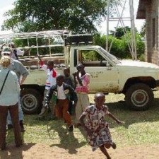 Kinder und Projektmitarbeiterin beim alten Geländewagen auf roter Erde