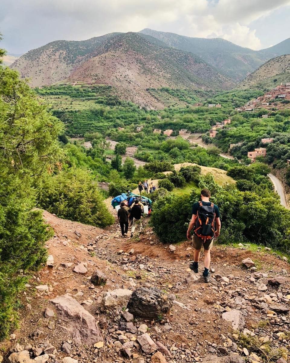 Trekkers hiking down mountain trail through lush green vegetation with valley villages