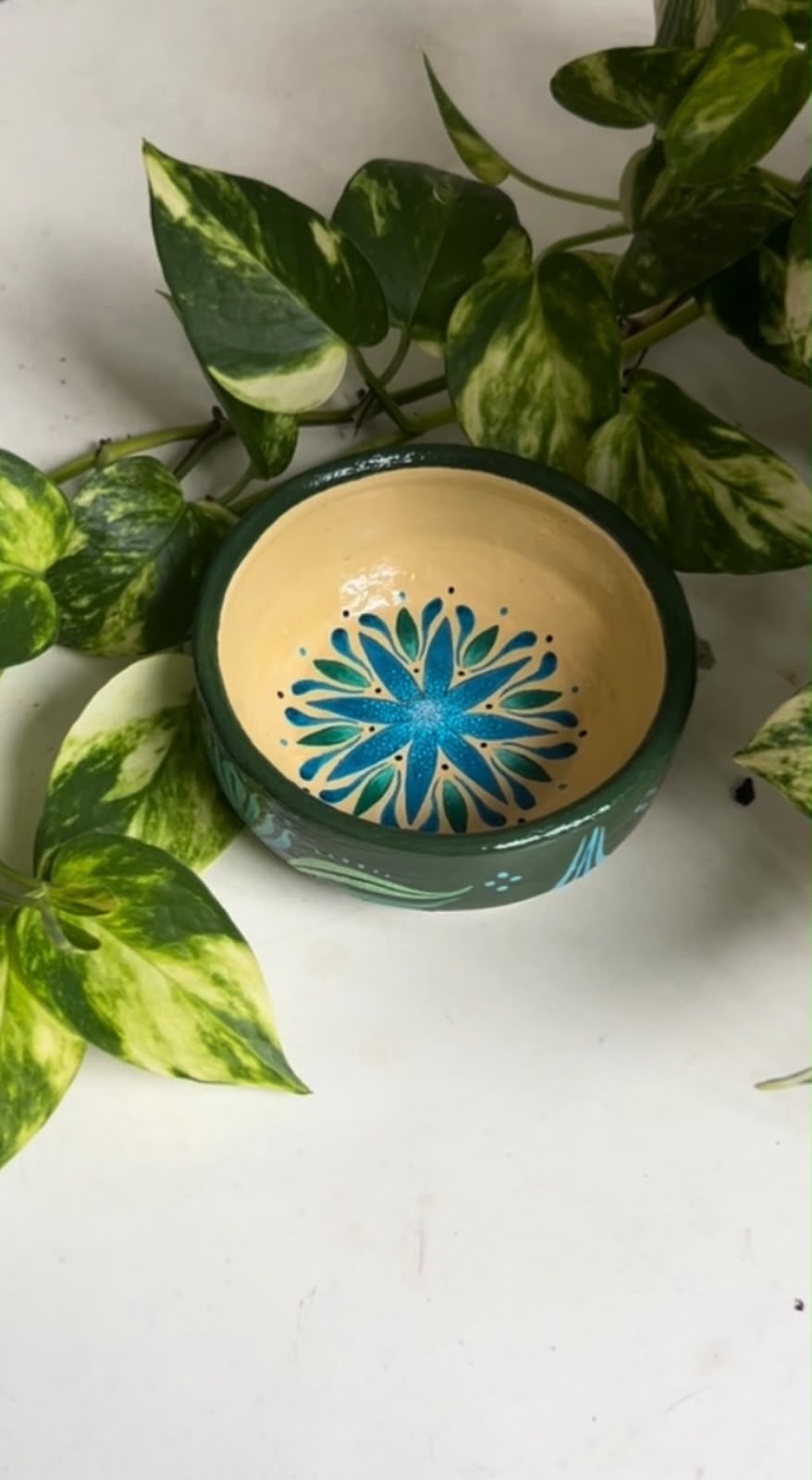 Potinho cerâmico verde com mandala azul e turquesa pintada à mão, visto de cima com folhagens