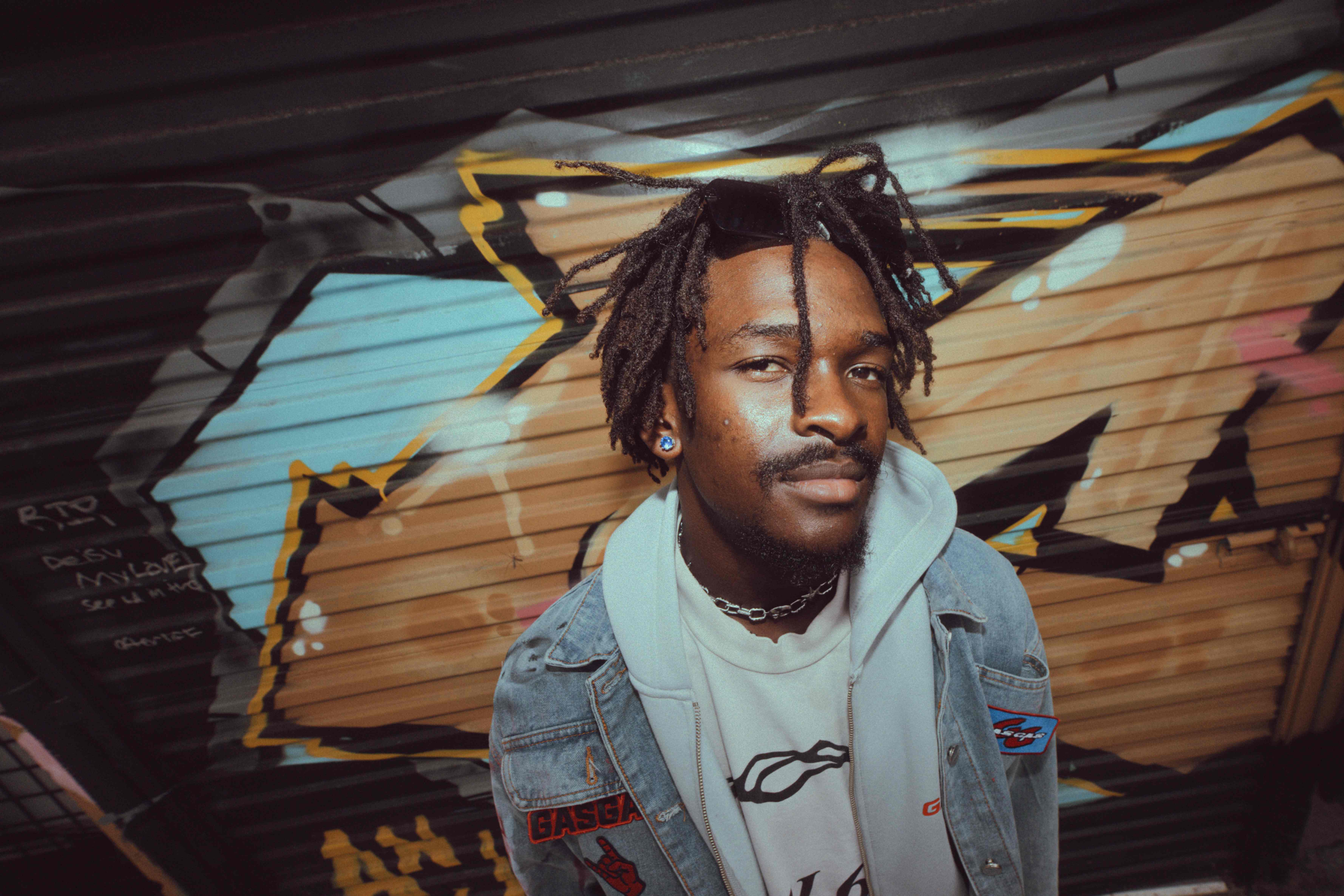 Man with dreadlocks in a denim GASGAS jacket posing against bright graffiti