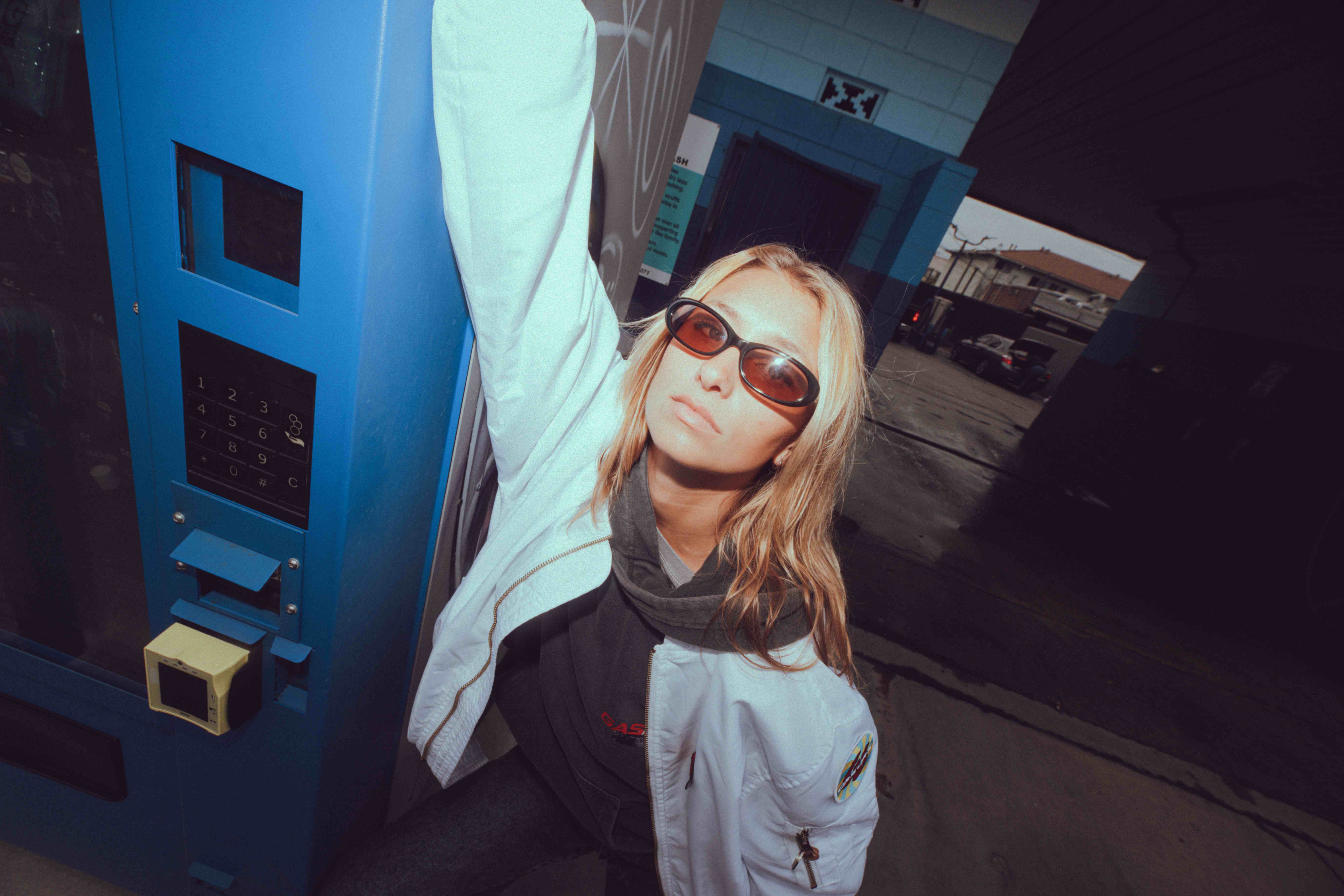 Blonde woman in a white bomber jacket and sunglasses leaning against a blue street machine