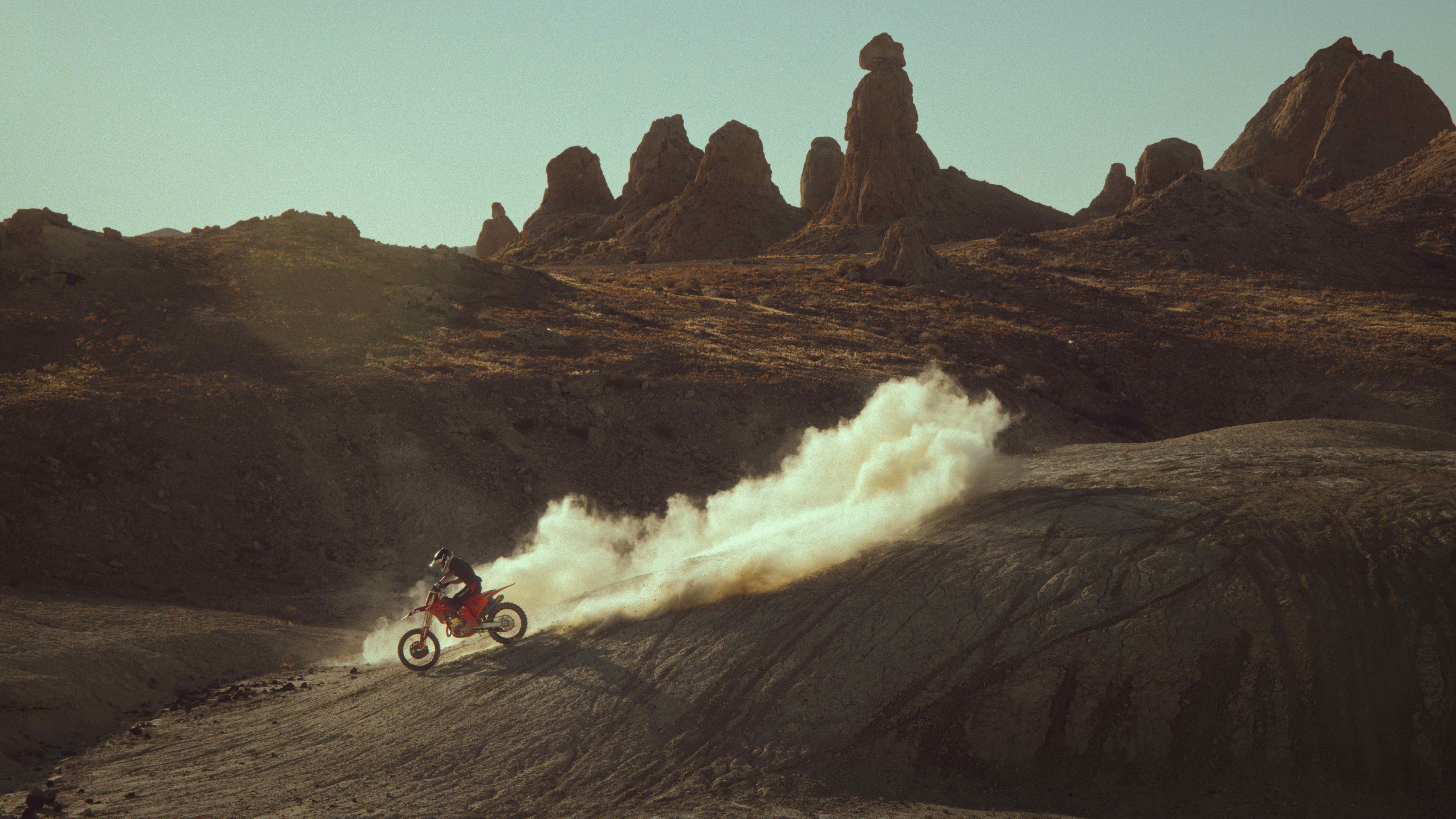 Enduro rider roosting a wall of dust beneath desert rock pinnacles at golden hour