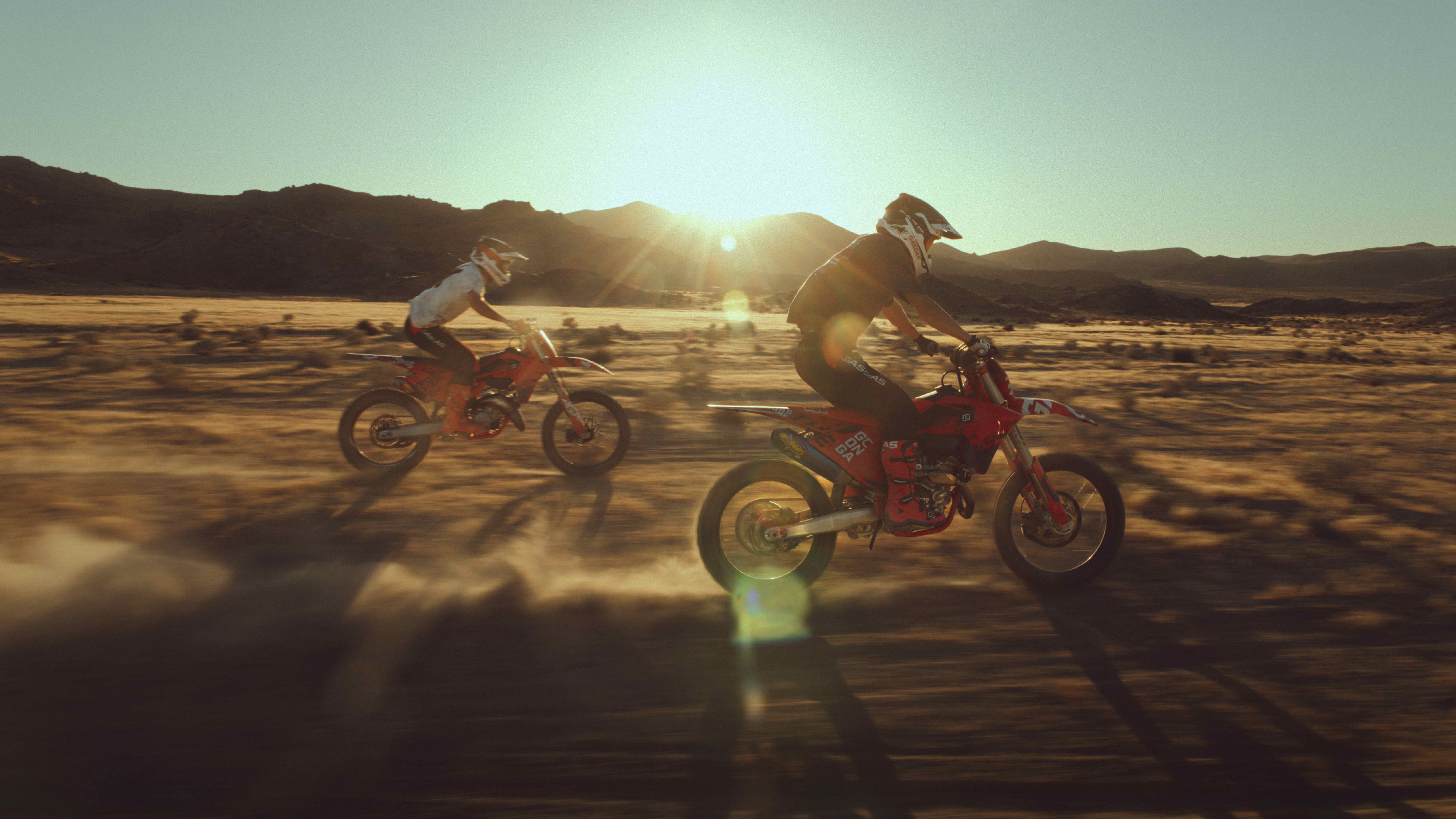 Two riders racing across desert flats into a low sun flare