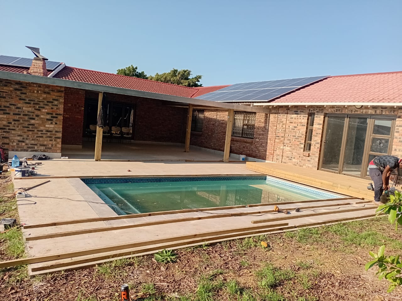 Before: Swimming pool timber deck Port Elizabeth — fresh pine boards being laid around pool, workers finishing edges, brick house with solar panels behind