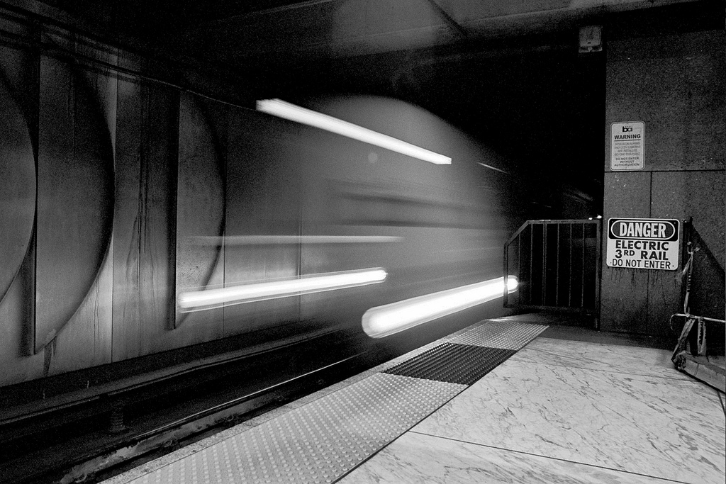 Underground transit platform with fluorescent lights, danger electrical signs, and motion-blurred arriving train