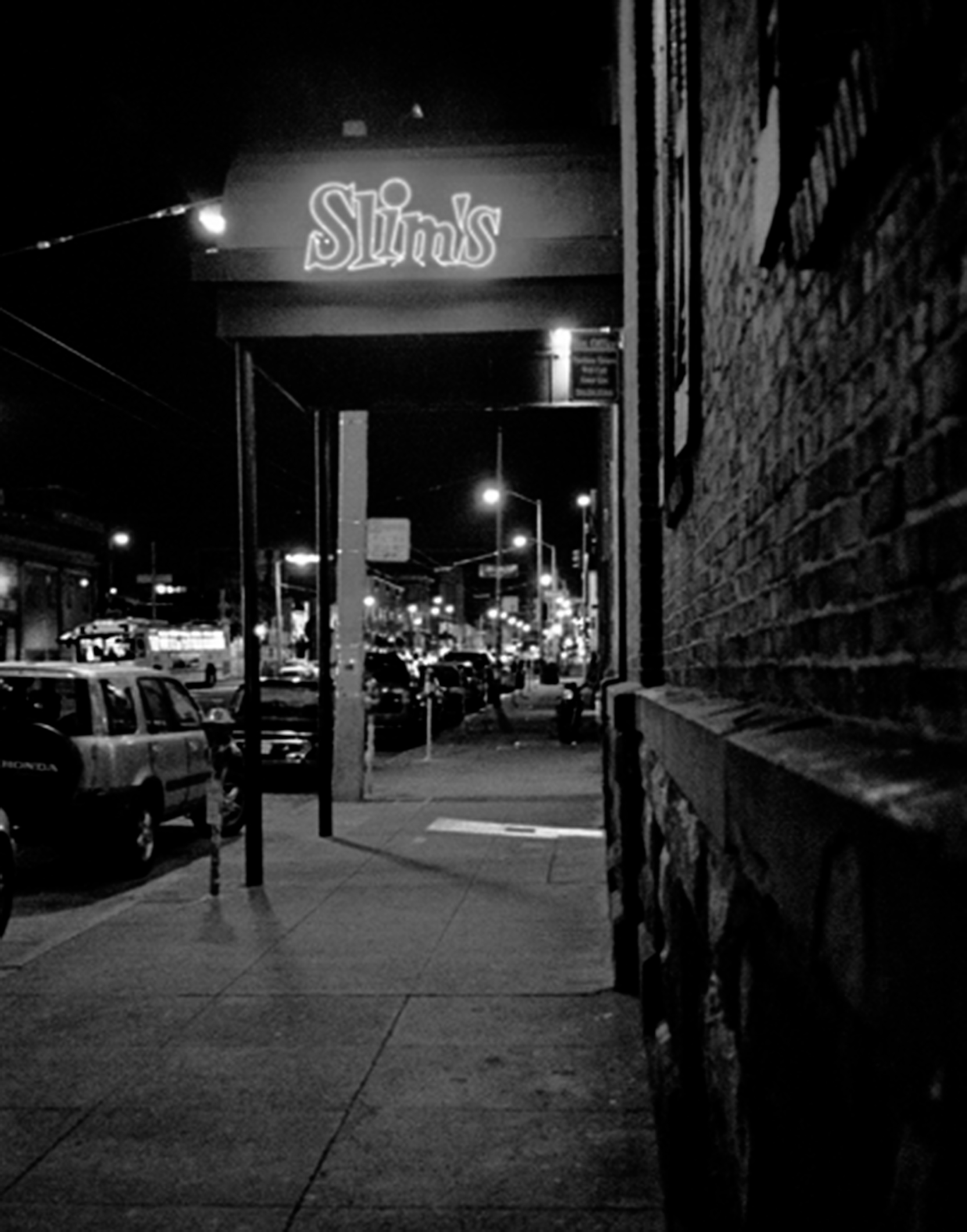 Neon sign spelling Slim's glowing at night with street cars, parked vehicles, and city lights blurred behind