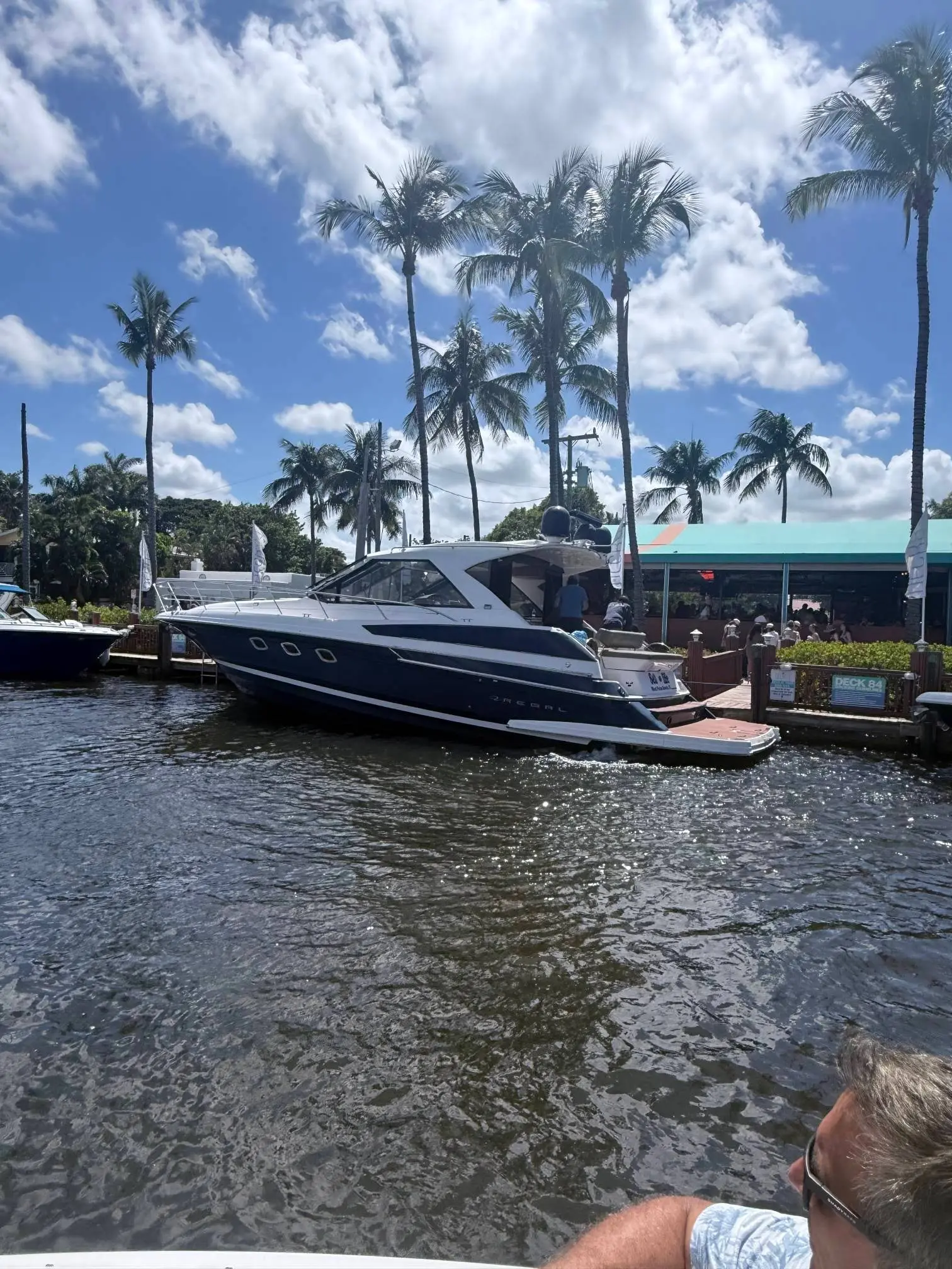 The Elite Sport Yachts 46ft Regal Sport Coupe cruising a Fort Lauderdale canal past Deck 84 restaurant, its navy blue hull gliding beneath tropical palm trees