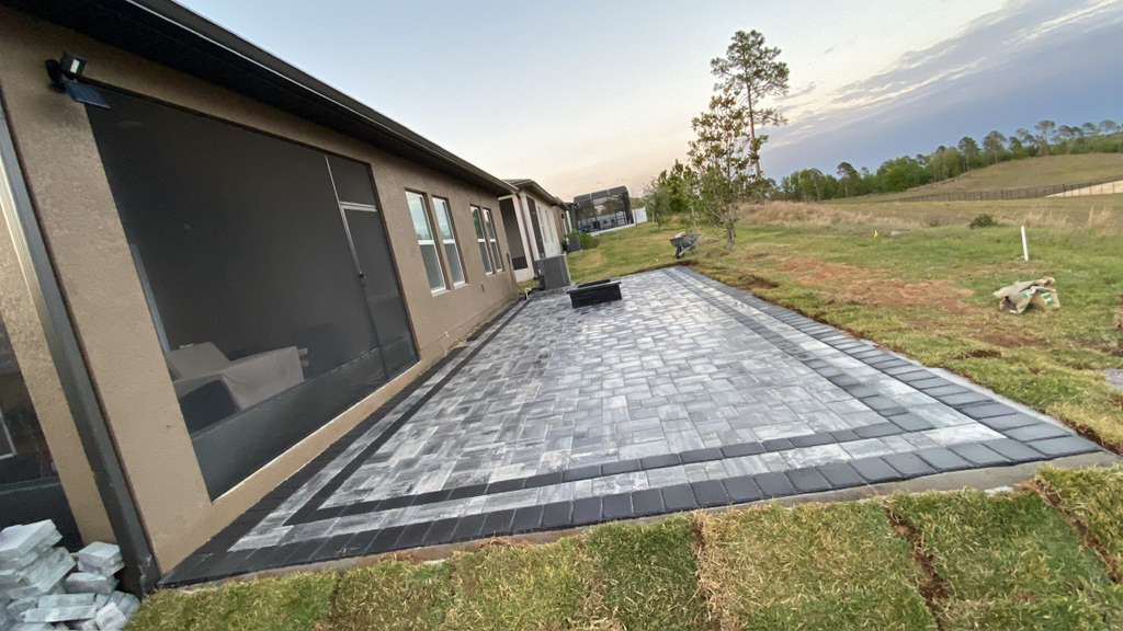 Large charcoal and grey brick paver patio along the side of a tan stucco home with contrasting dark border edging at dusk