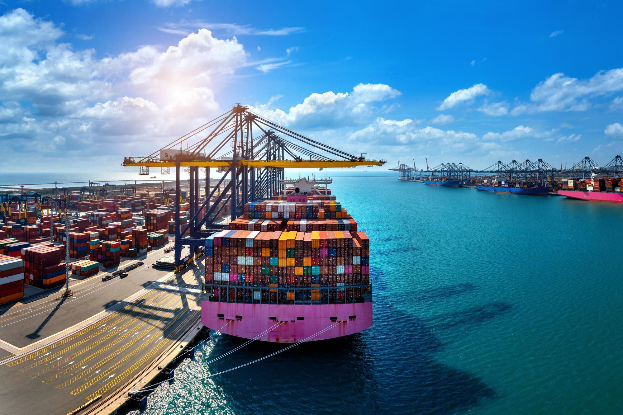 Aerial view of a container port showing a large container ship with pink hull loaded with colorful shipping containers, alongside gantry cranes and turquoise ocean waters, symbolizing international trade and supply chain operations