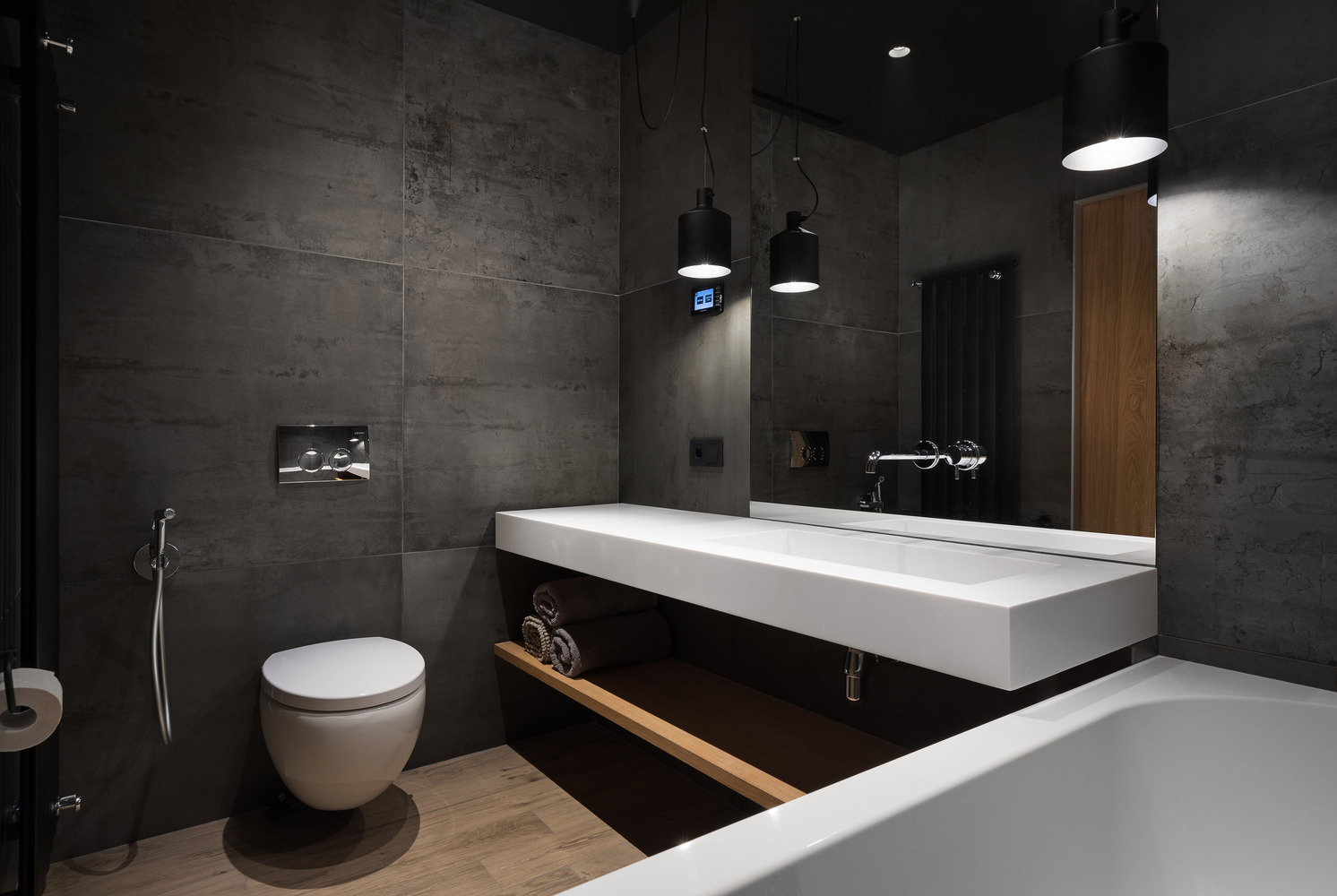 Industrial dark bathroom with large-format concrete-look tiles, floating white trough sink with open wood shelf, black pendant lights, wall-hung toilet, chrome fixtures, wood-plank floor