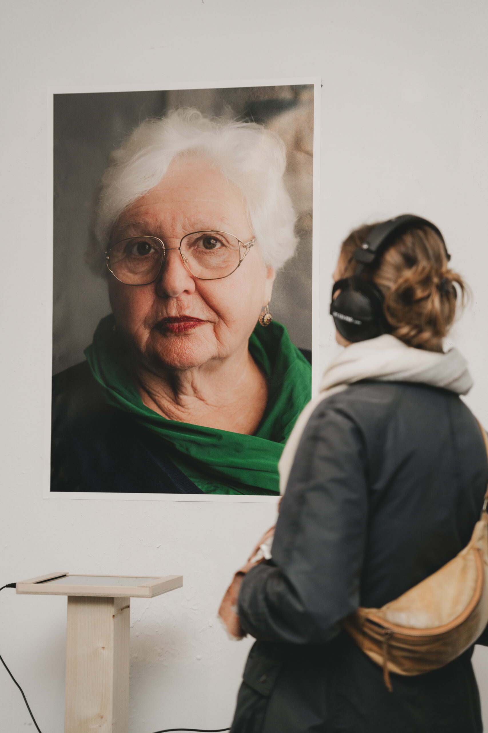 Ausstellungsbesucherin mit Kopfhörern betrachtet ein großformatiges Porträt einer älteren Frau