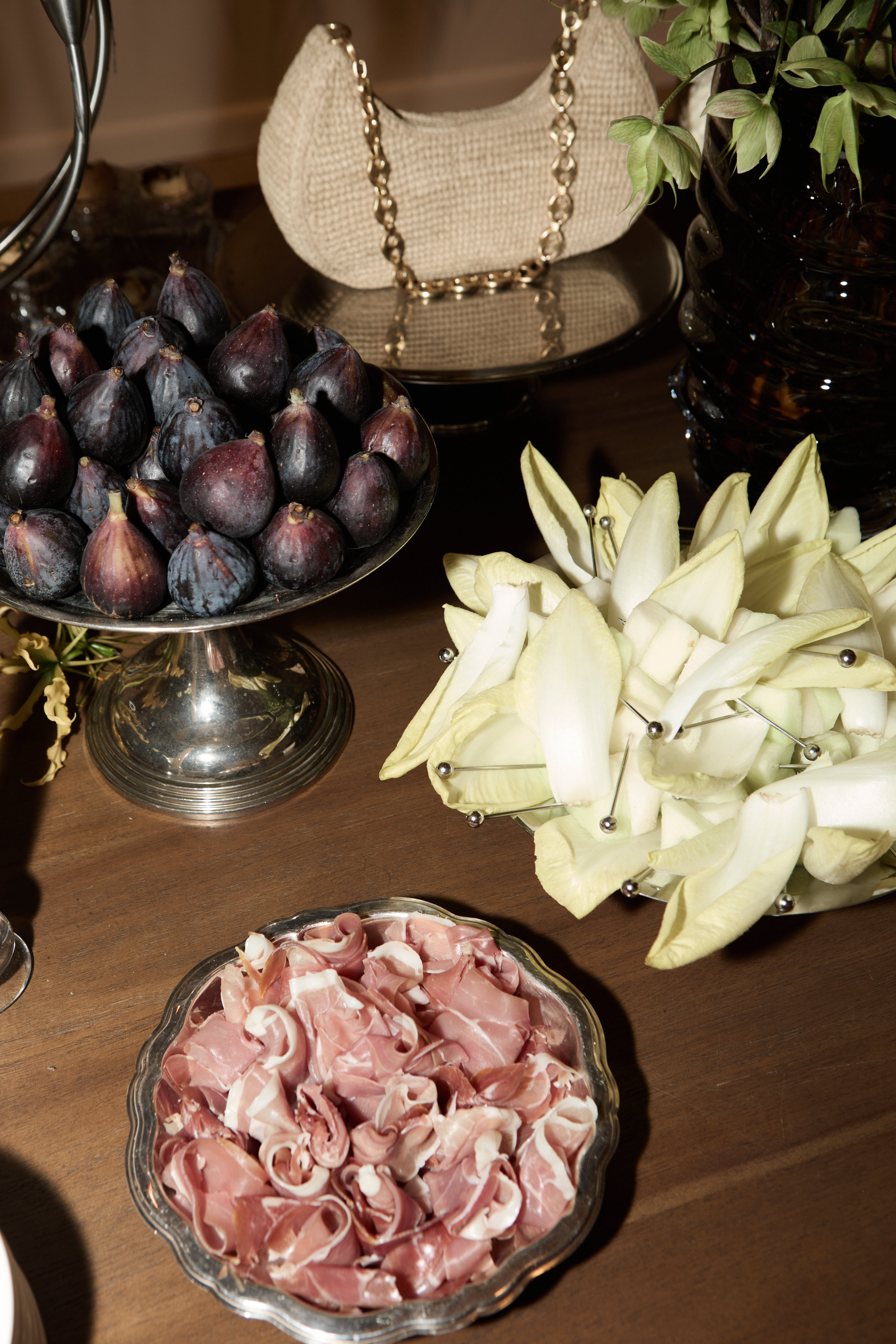 Grazing table spread with black figs, endive, prosciutto, and a raffia bag