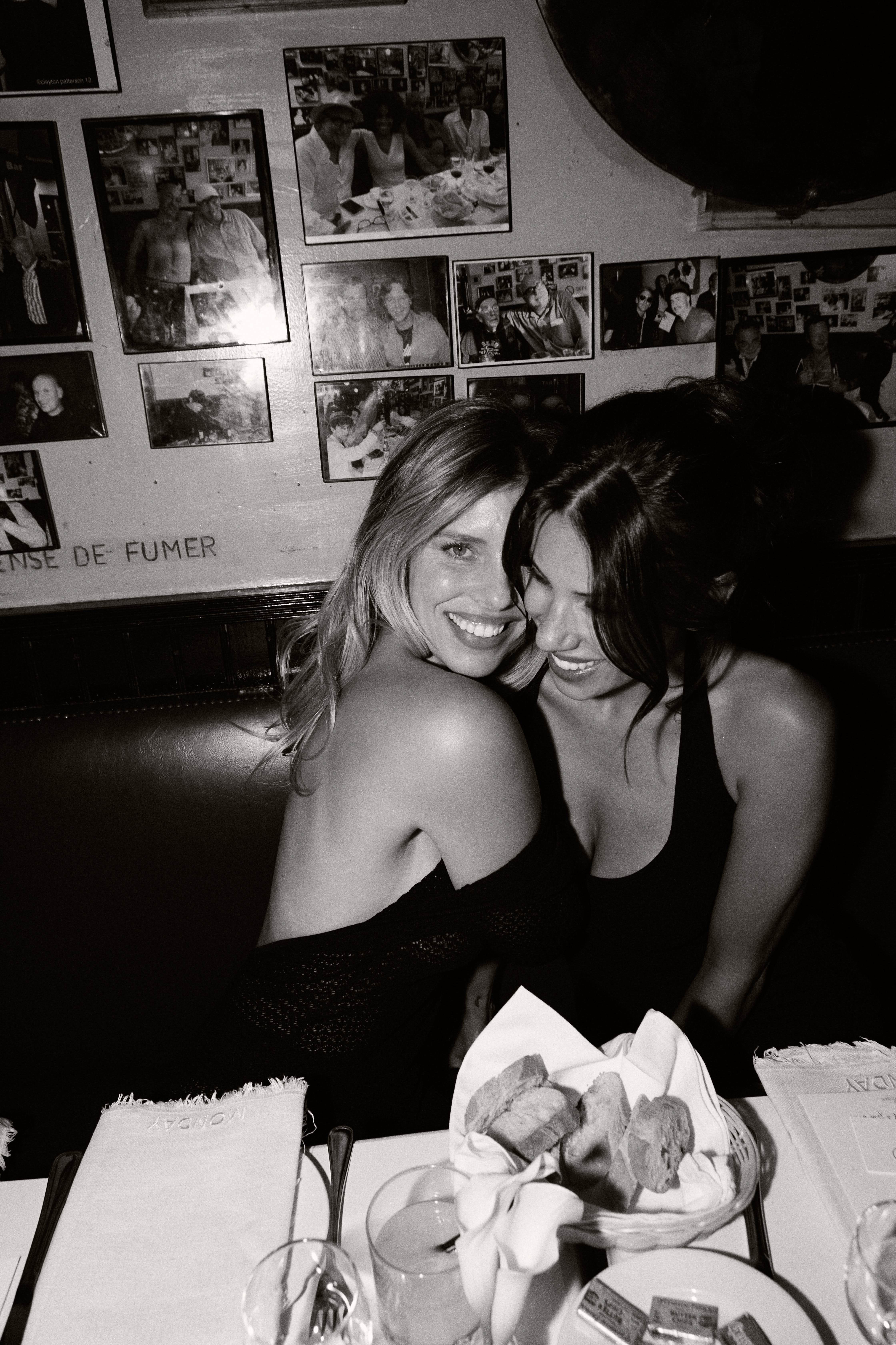 Black and white shot of two smiling women embracing in a photo-covered restaurant booth