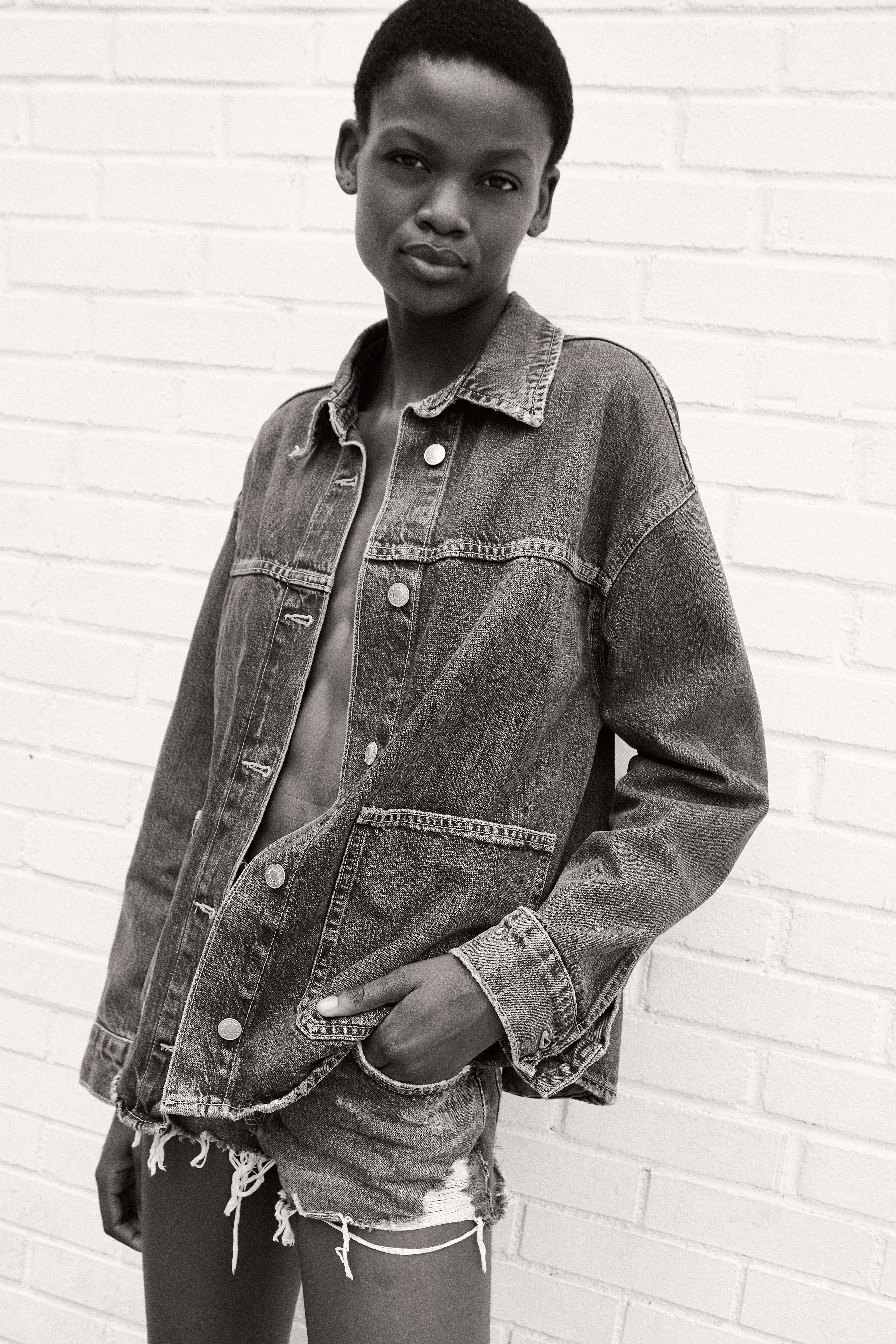 Black and white model in an oversized washed denim jacket and cut-off shorts against a white brick wall