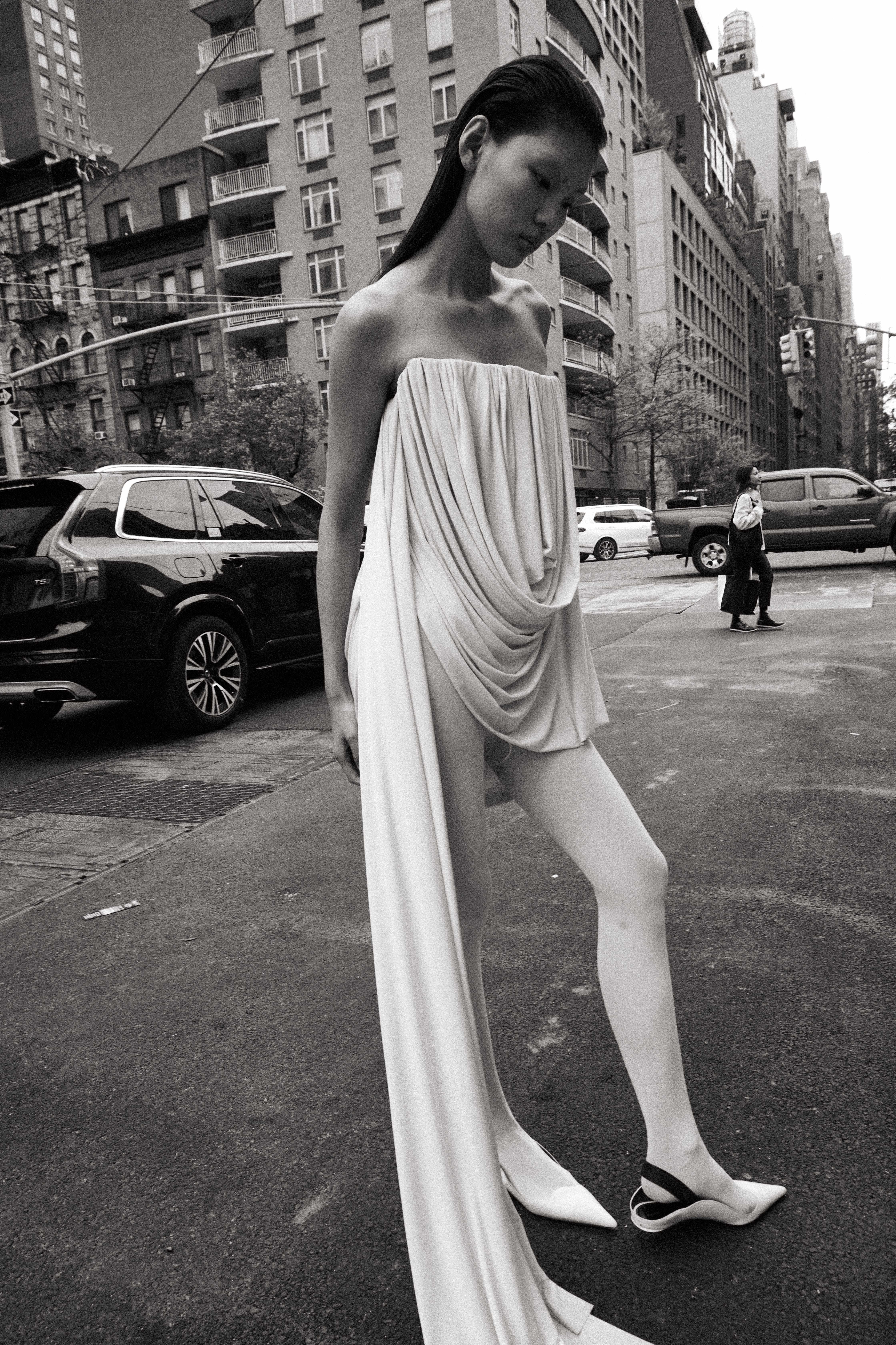 Black and white street shot of model in draped strapless white gown walking NYC street