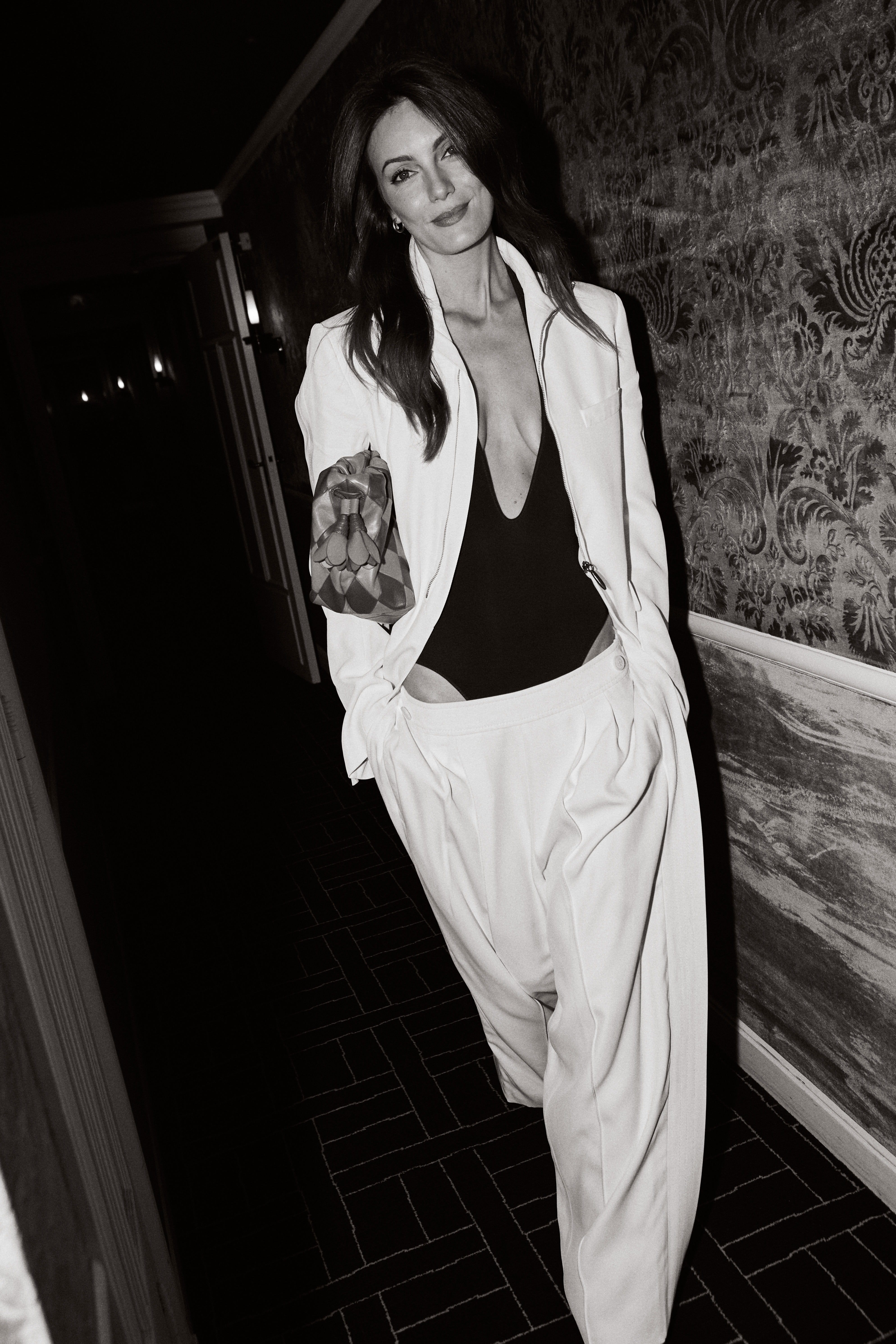 Woman in white oversized blazer suit with black plunging top holding patterned clutch in elegant hallway