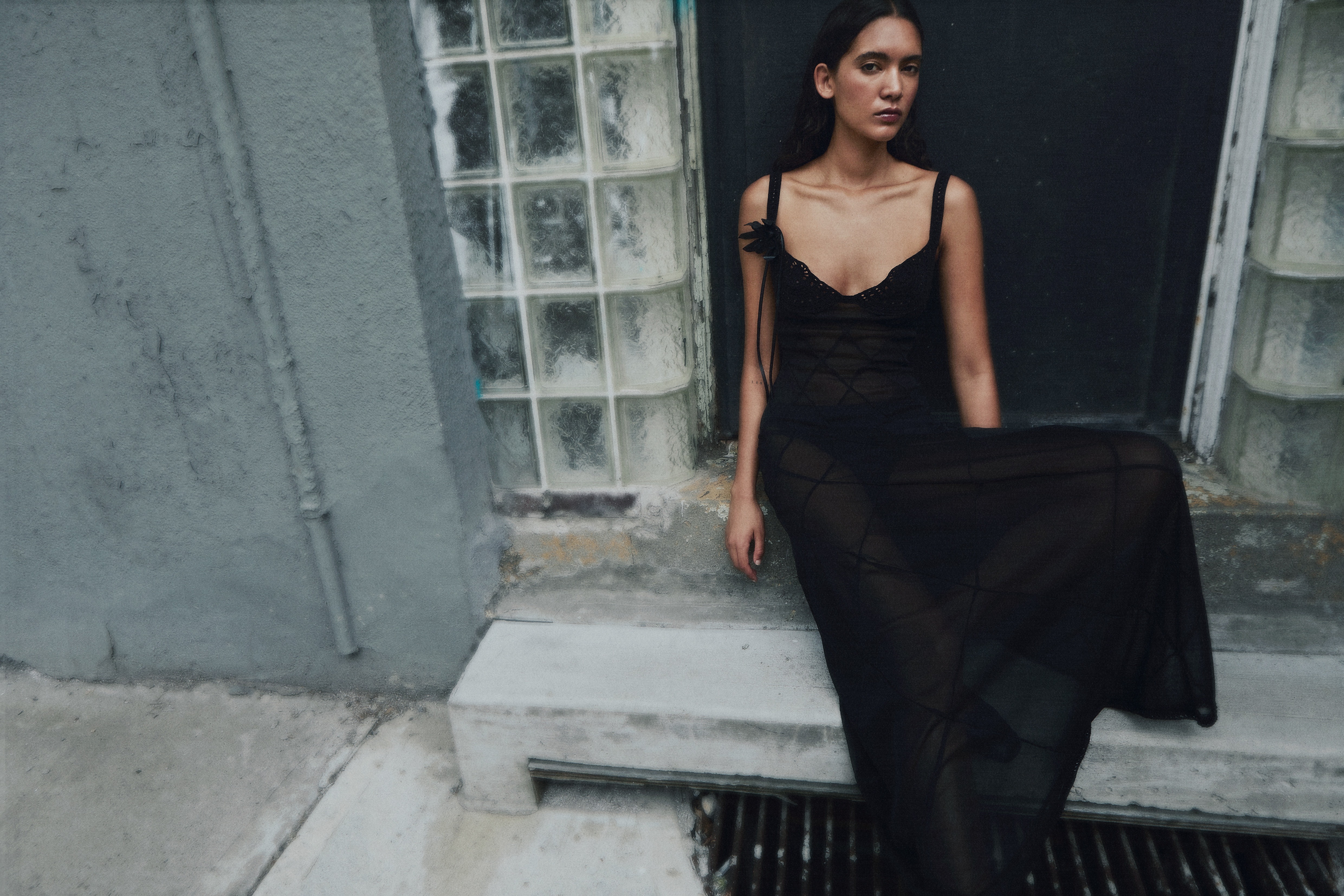 Woman in sheer black slip dress leaning against weathered concrete wall with glass block window