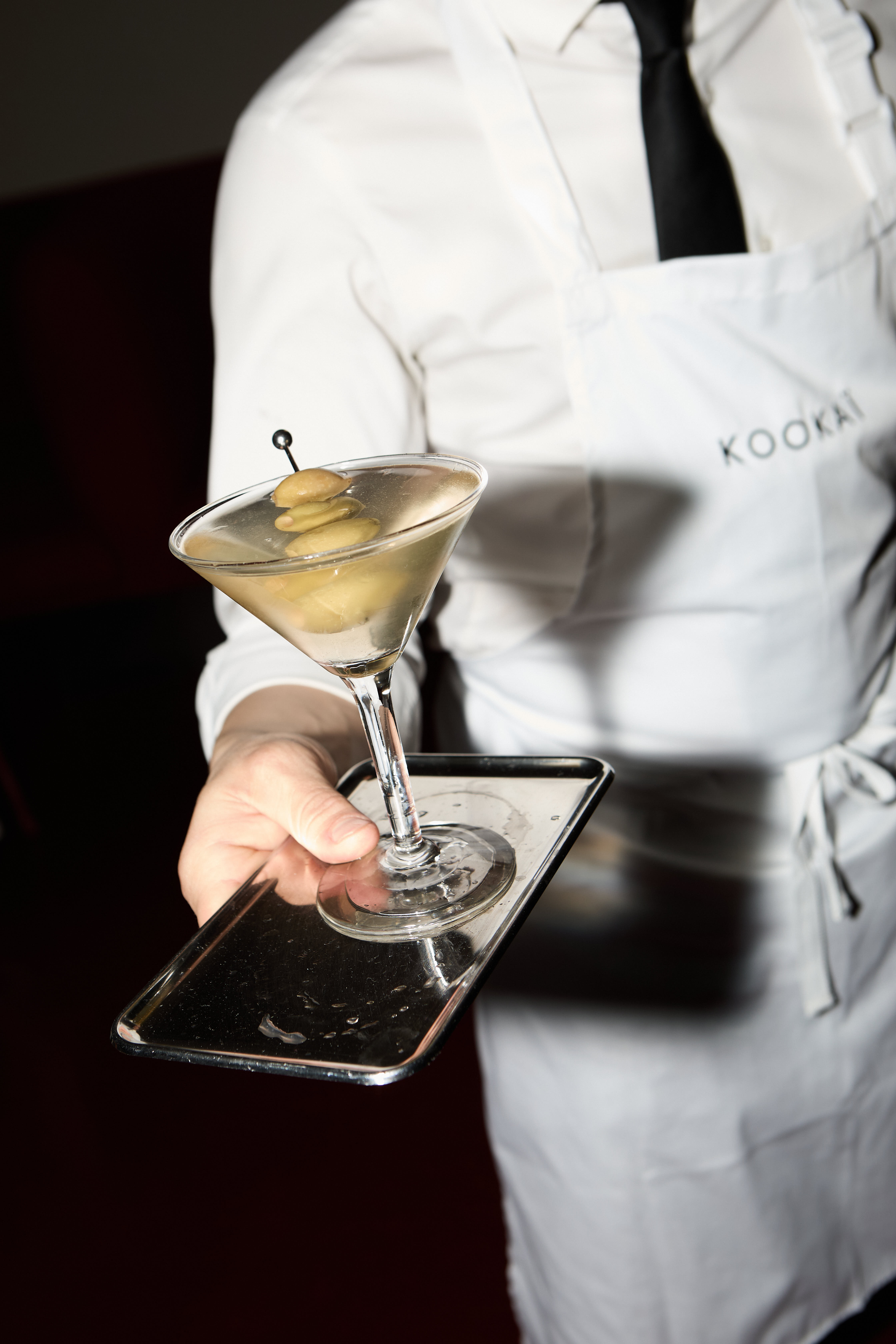 Waiter in white apron holding a dirty martini on a silver tray