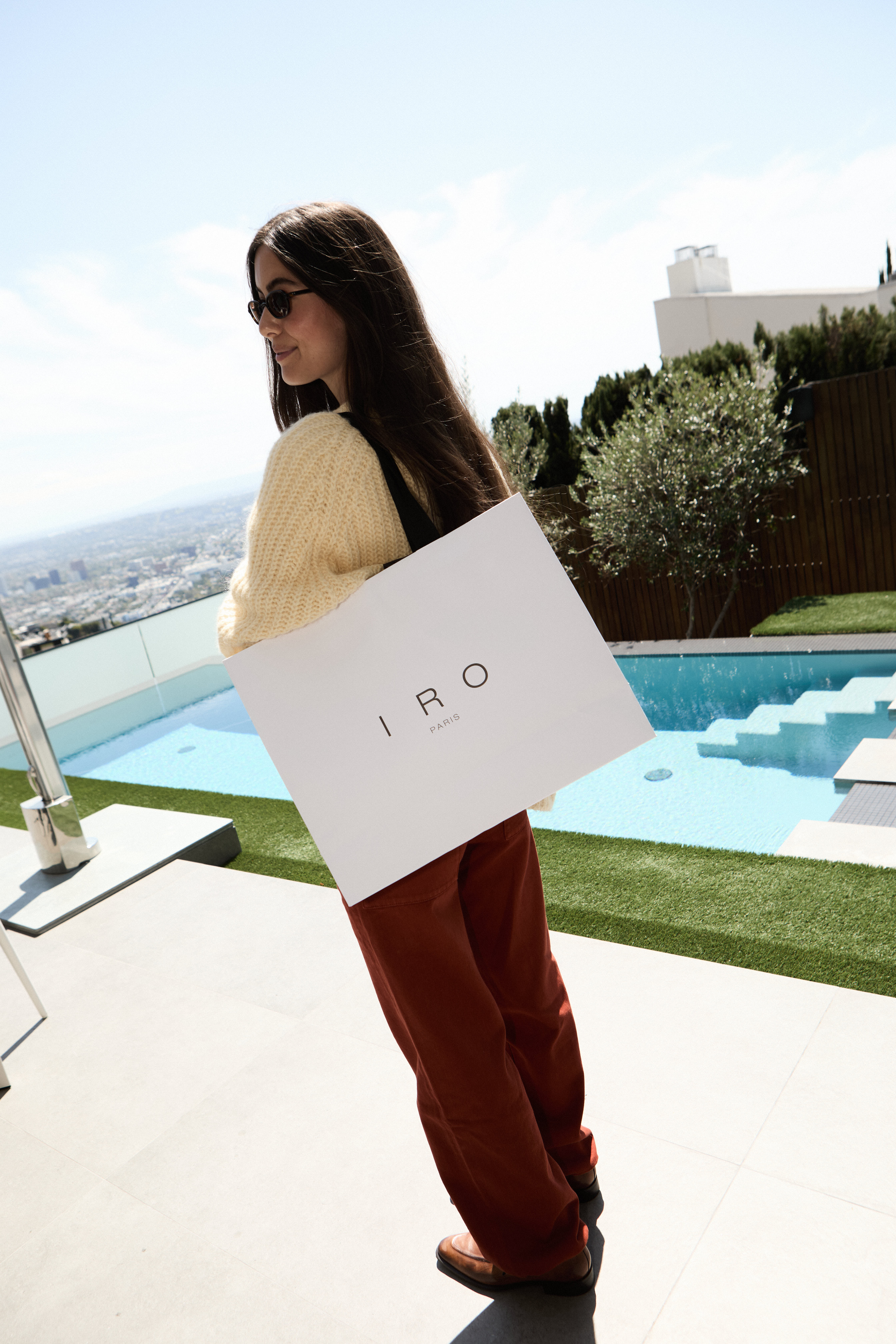 Woman in cream sweater and rust trousers holding a white IRO Paris bag by a hillside pool