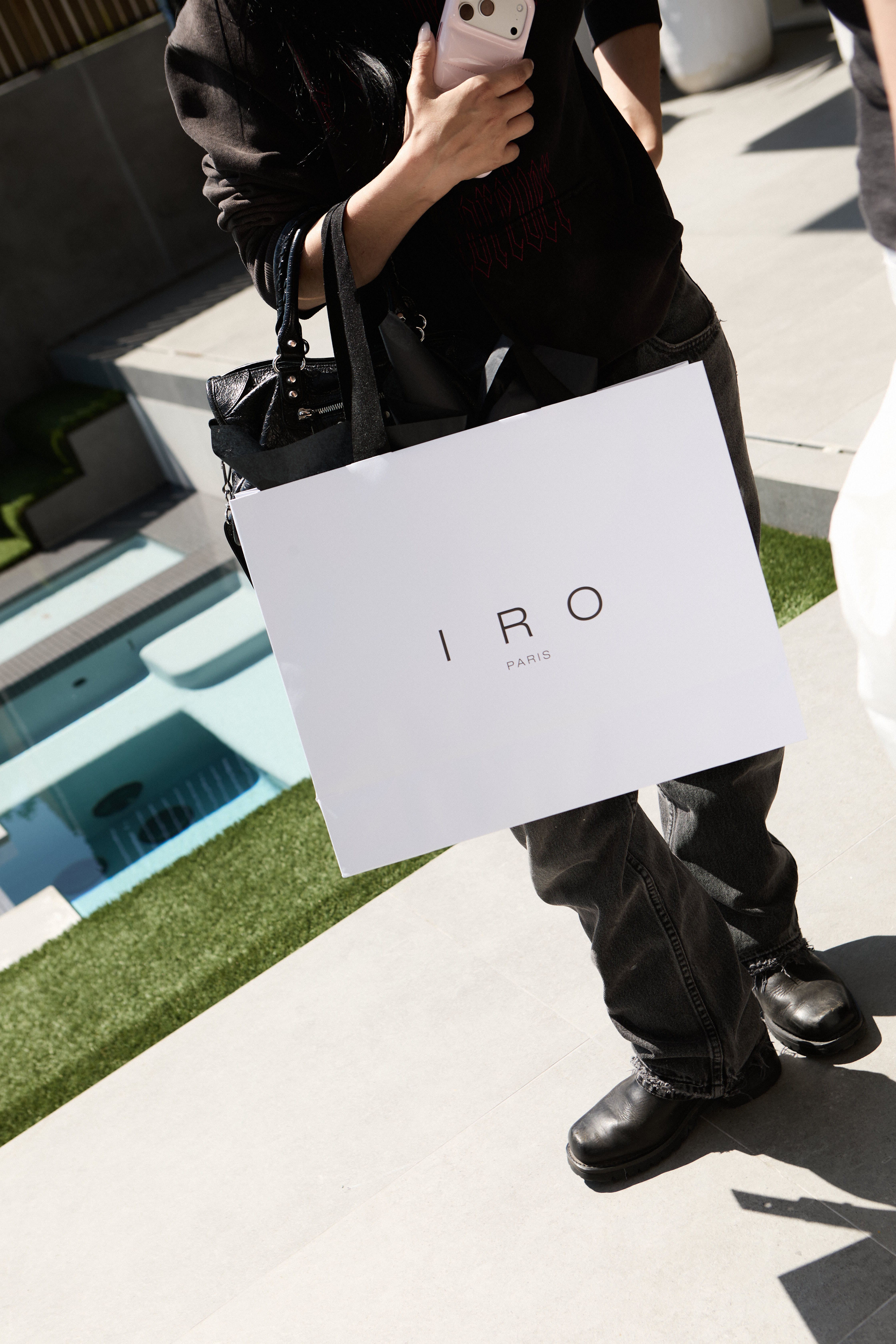 Person in black holding a white IRO Paris shopping bag by a pool