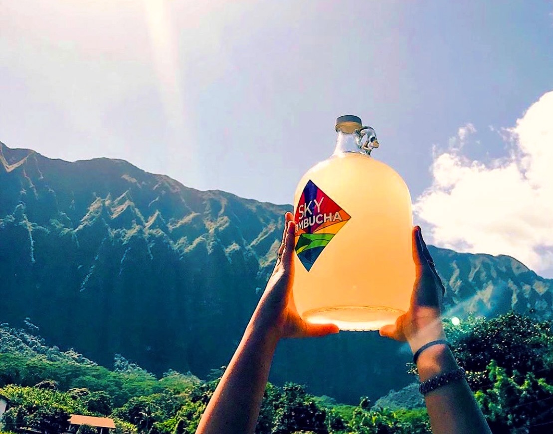 SKY Kombucha growler held up against Ko'olau mountains with sun flare