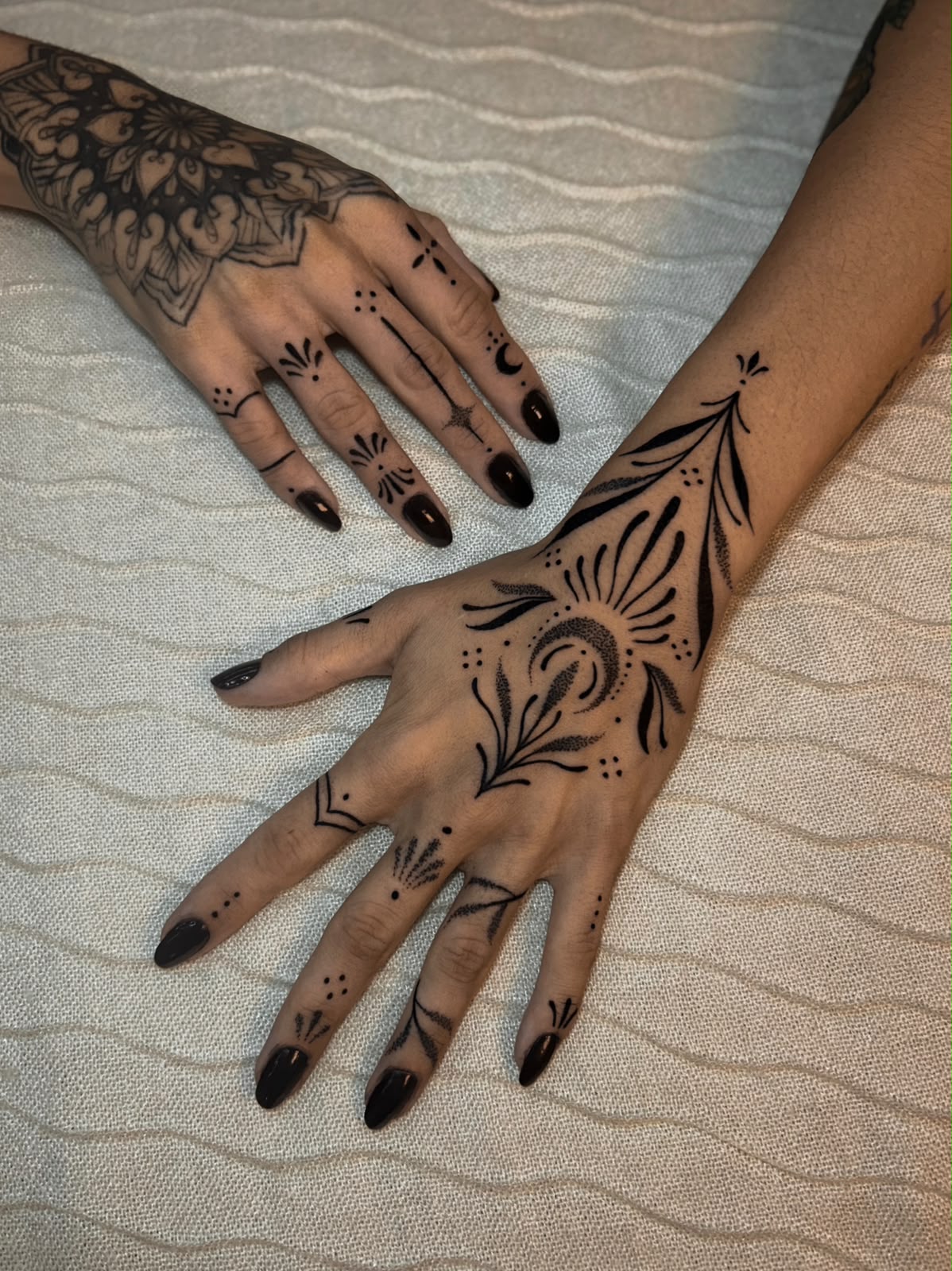Duas mãos com tatuagens ornamentais nos dedos e dorso, unhas pretas
