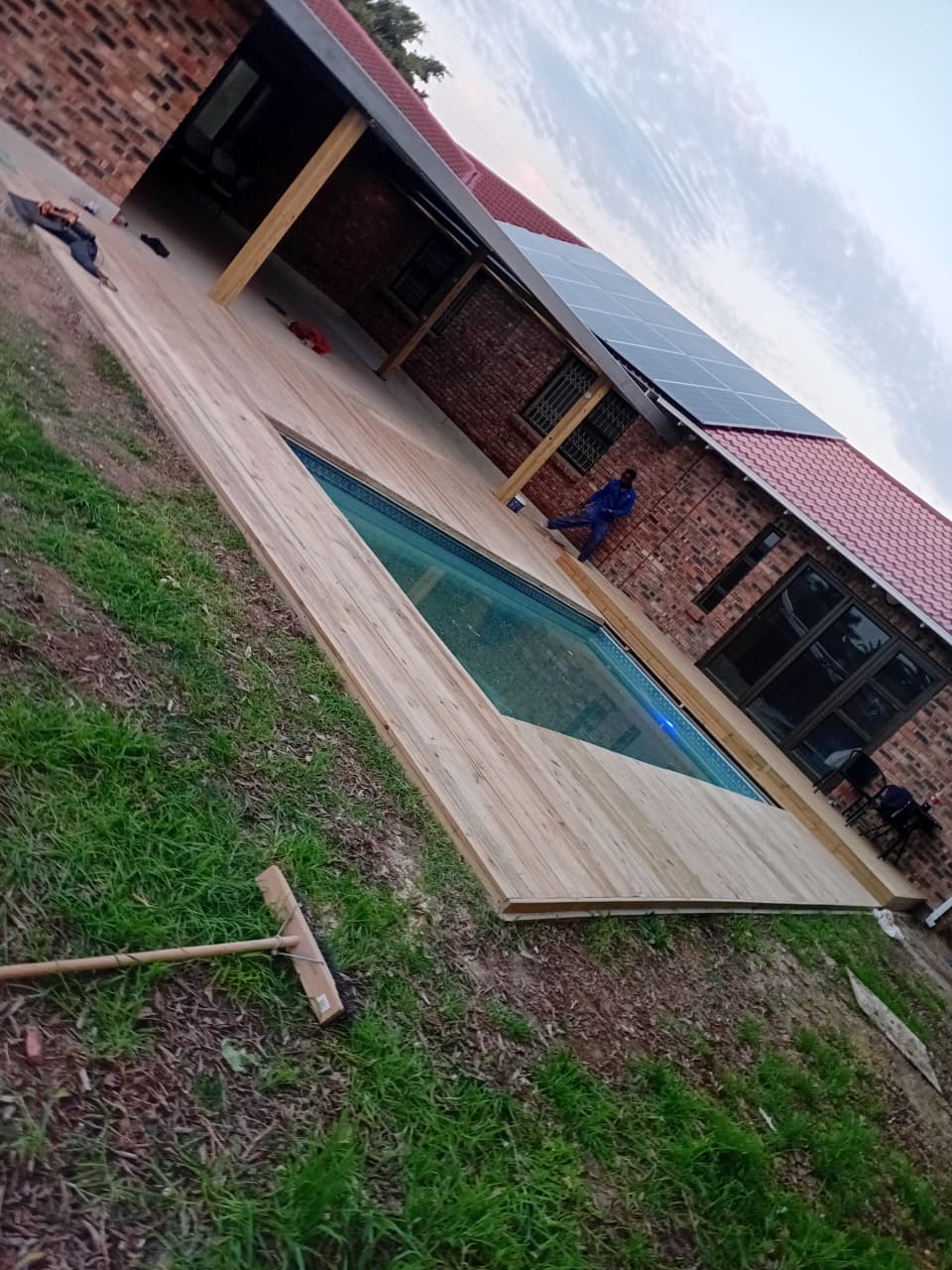 After: Completed swimming pool wooden deck Port Elizabeth — full wrap-around natural pine deck surrounding pool, brick house with solar panels, pergola posts