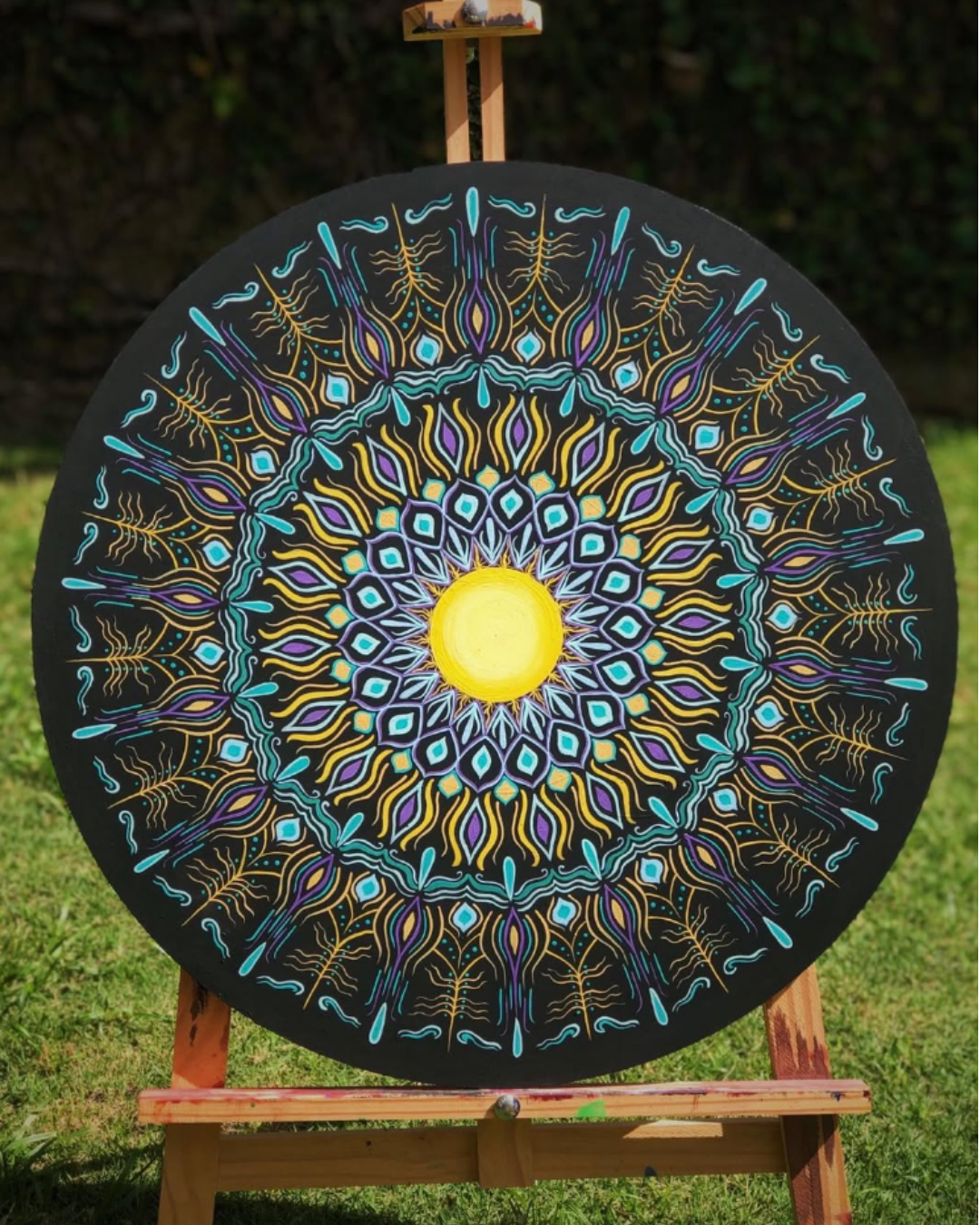 Mandala circular preta com padrão em turquesa, roxo e amarelo dourado sobre cavalete na grama