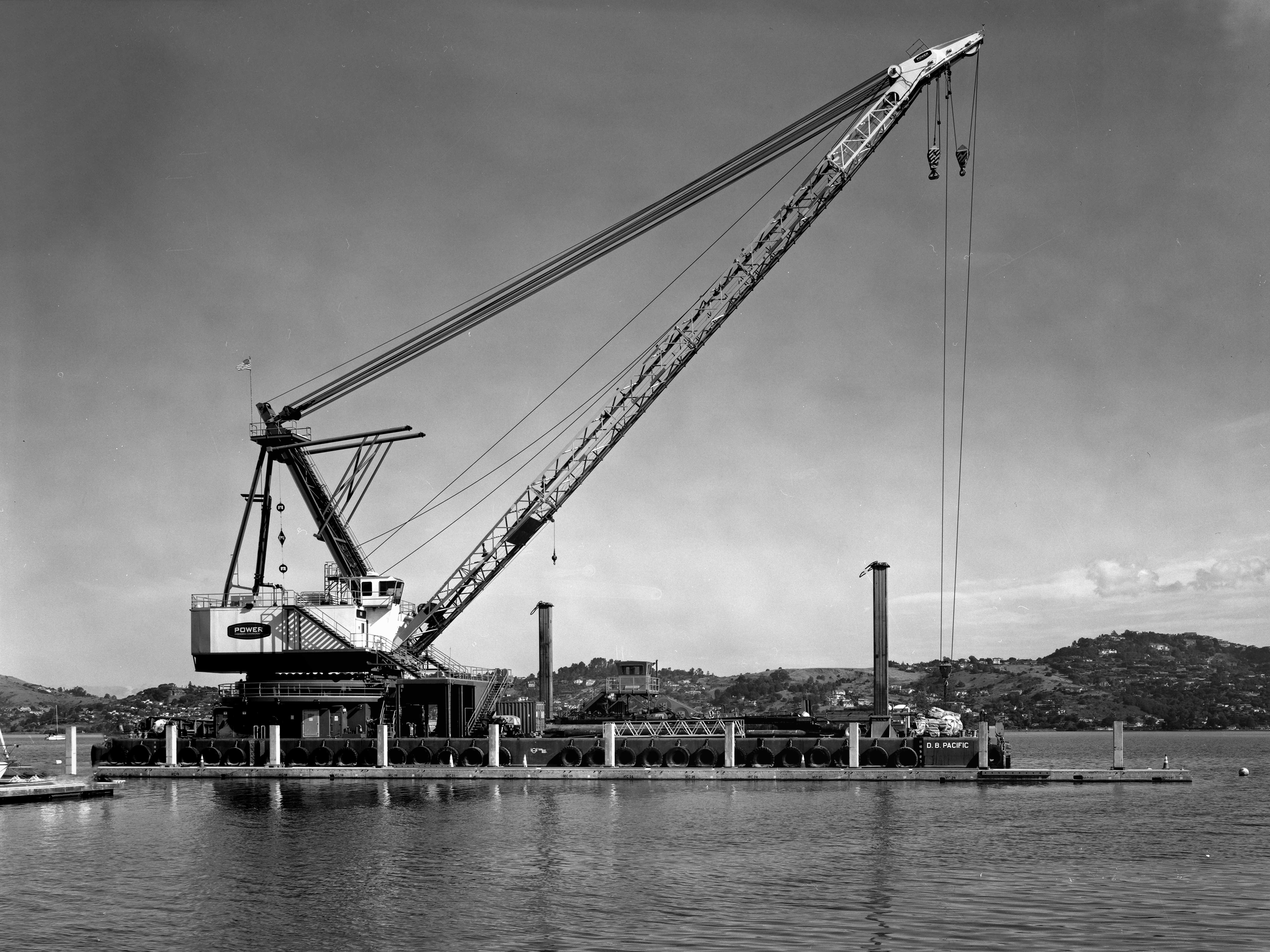 Large industrial cargo crane at waterfront dock with extended boom arm, cables, and hillside landscape in background