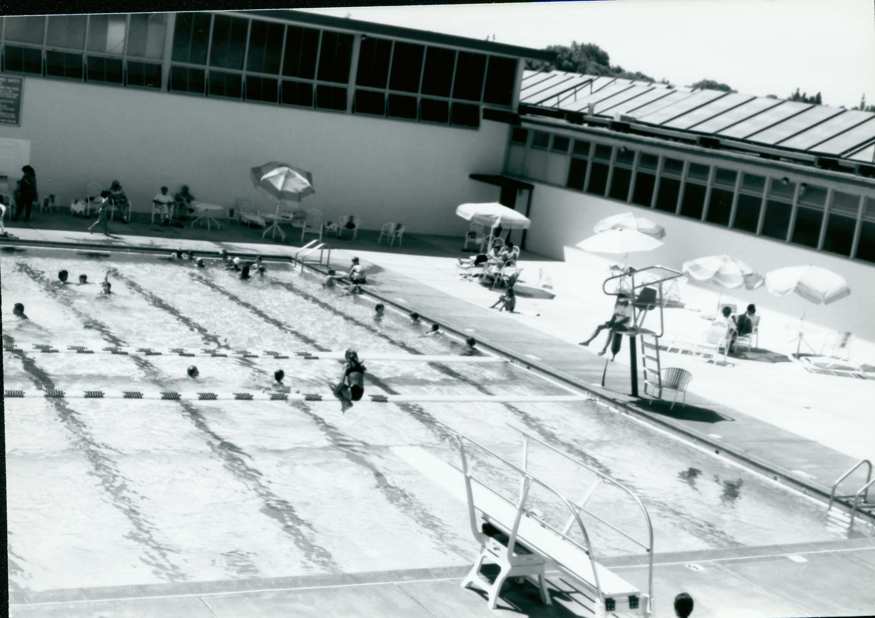 A high view across the Campbell pool showing swimmers, lane lines, umbrellas, and the surrounding building.