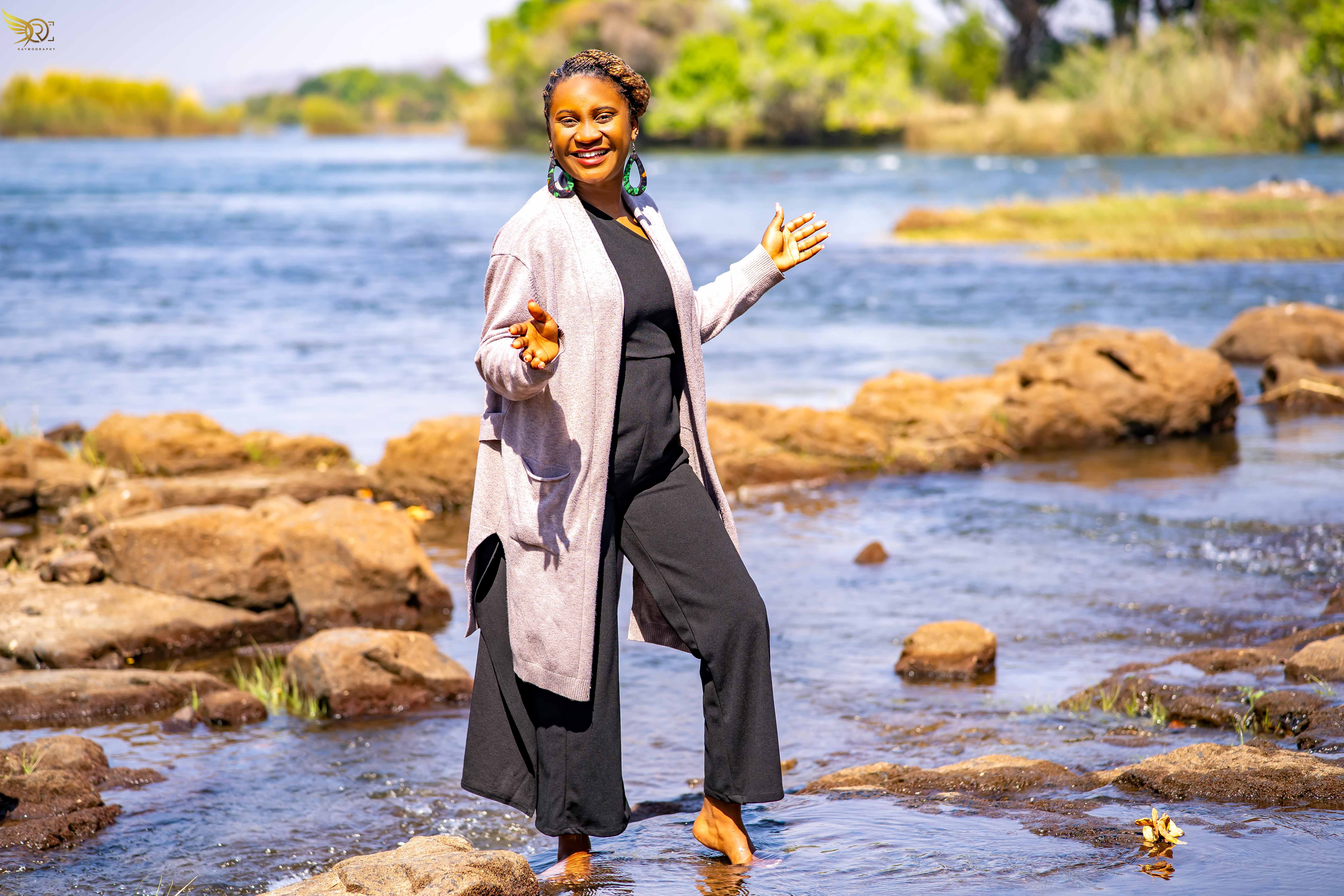 Tendai Makoni at riverbank wearing cream cardigan and black outfit in joyful pose
