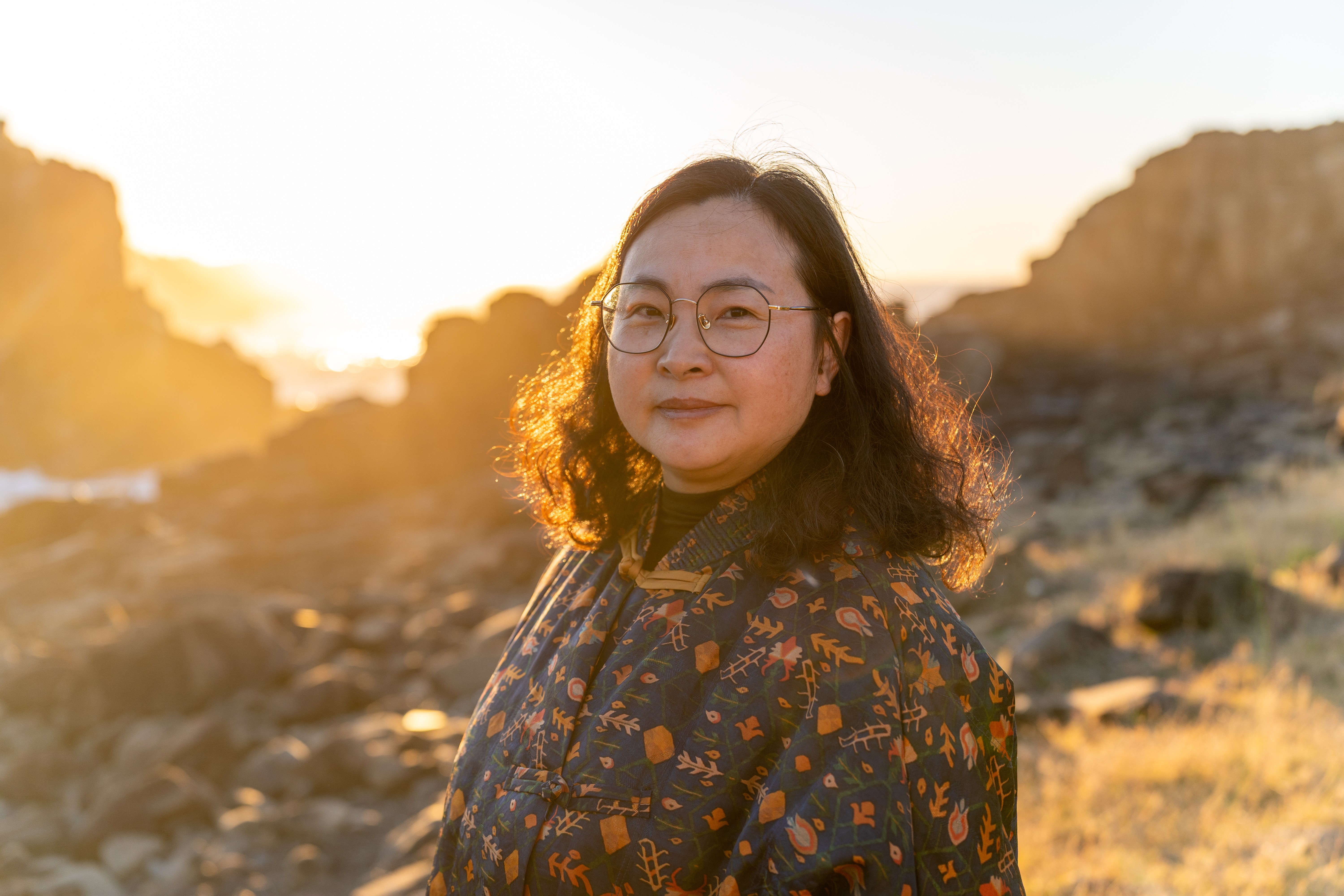 Woman in glasses backlit by a golden sunrise among coastal rocks