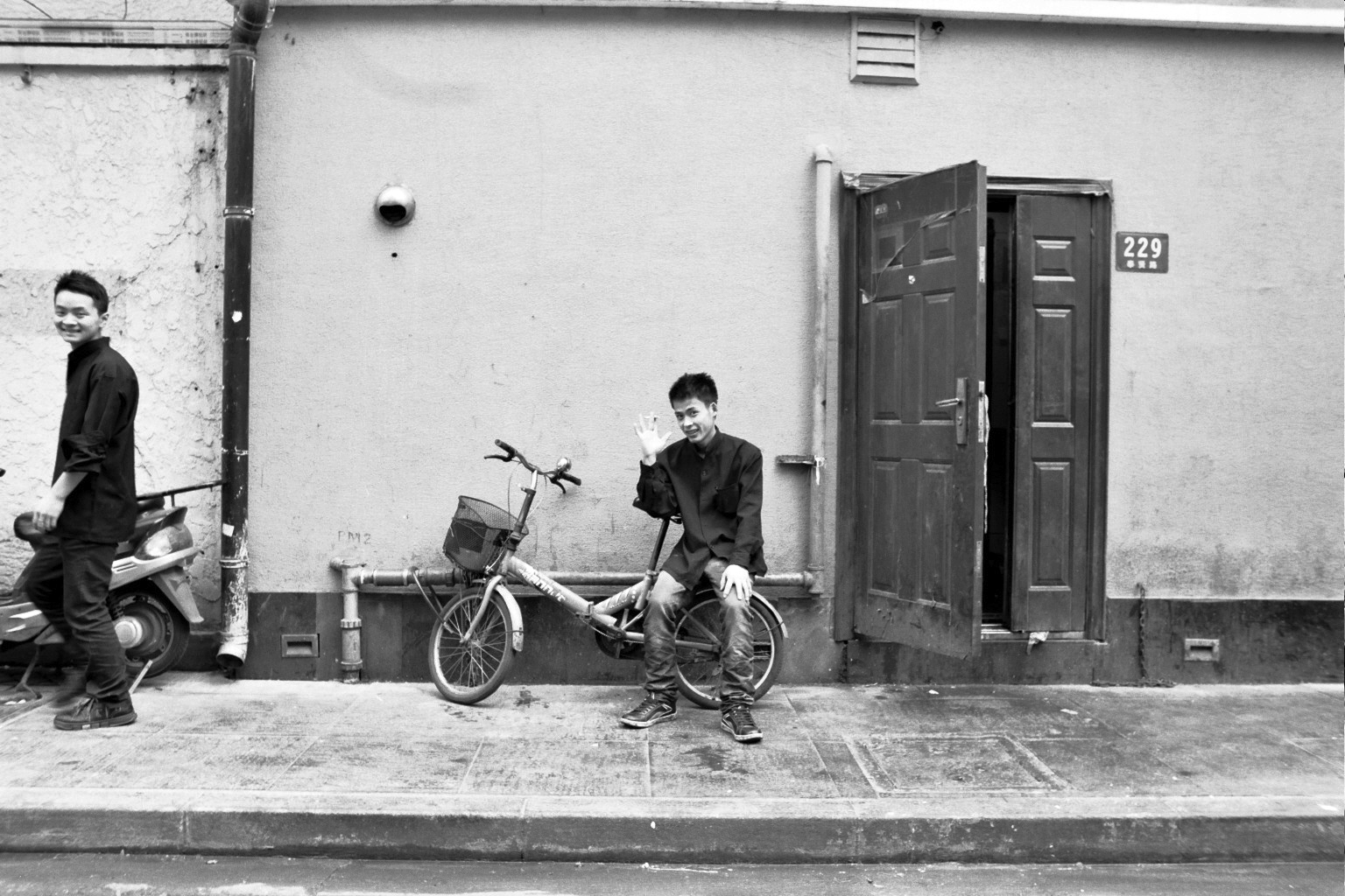 Young man sitting on bicycle waving at camera on urban street with door number 229