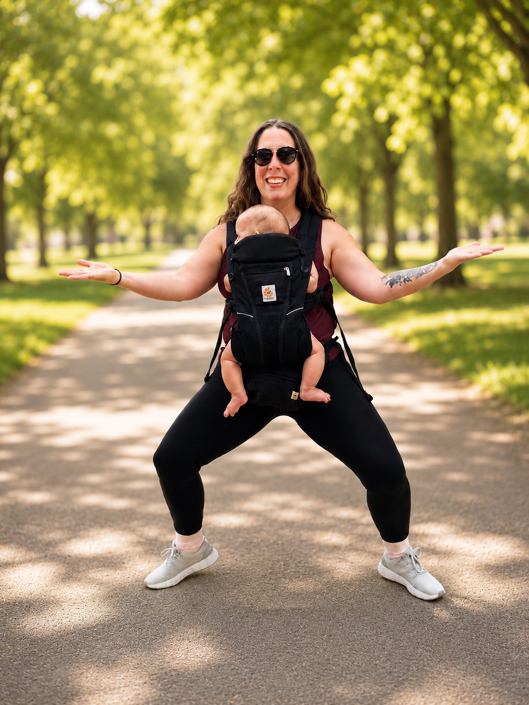 The instructor in a wide squat stance outdoors with baby in carrier, arms spread wide and smiling