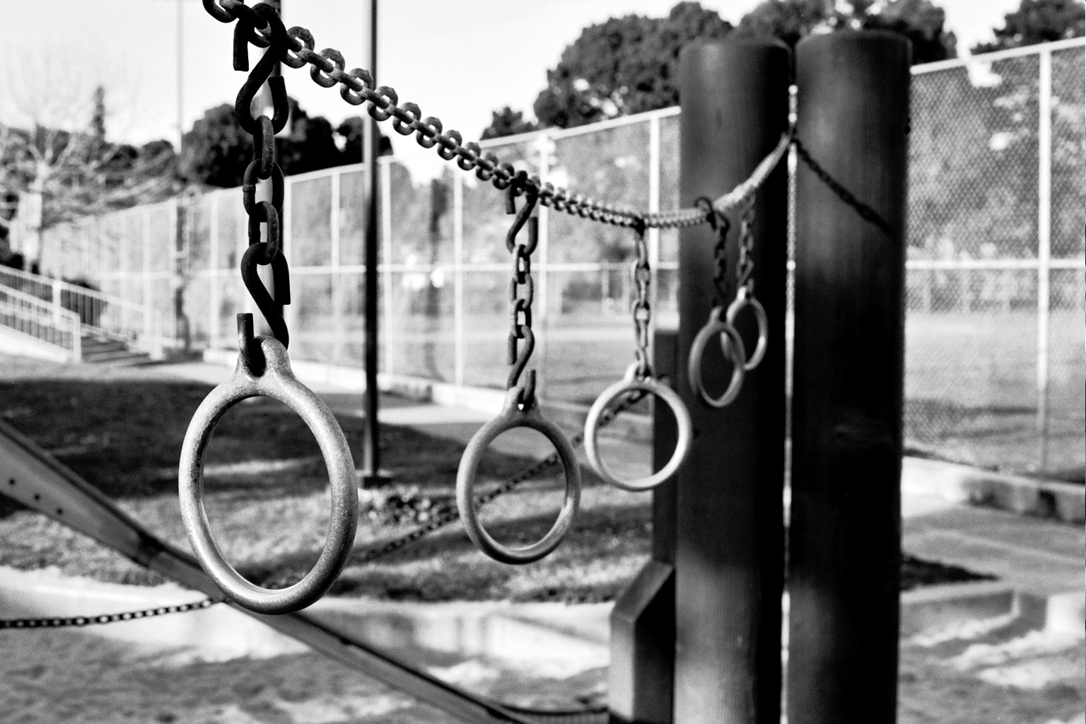 Close-up of metal playground rings hanging from chain with gymnasium and chain link fence blurred in background
