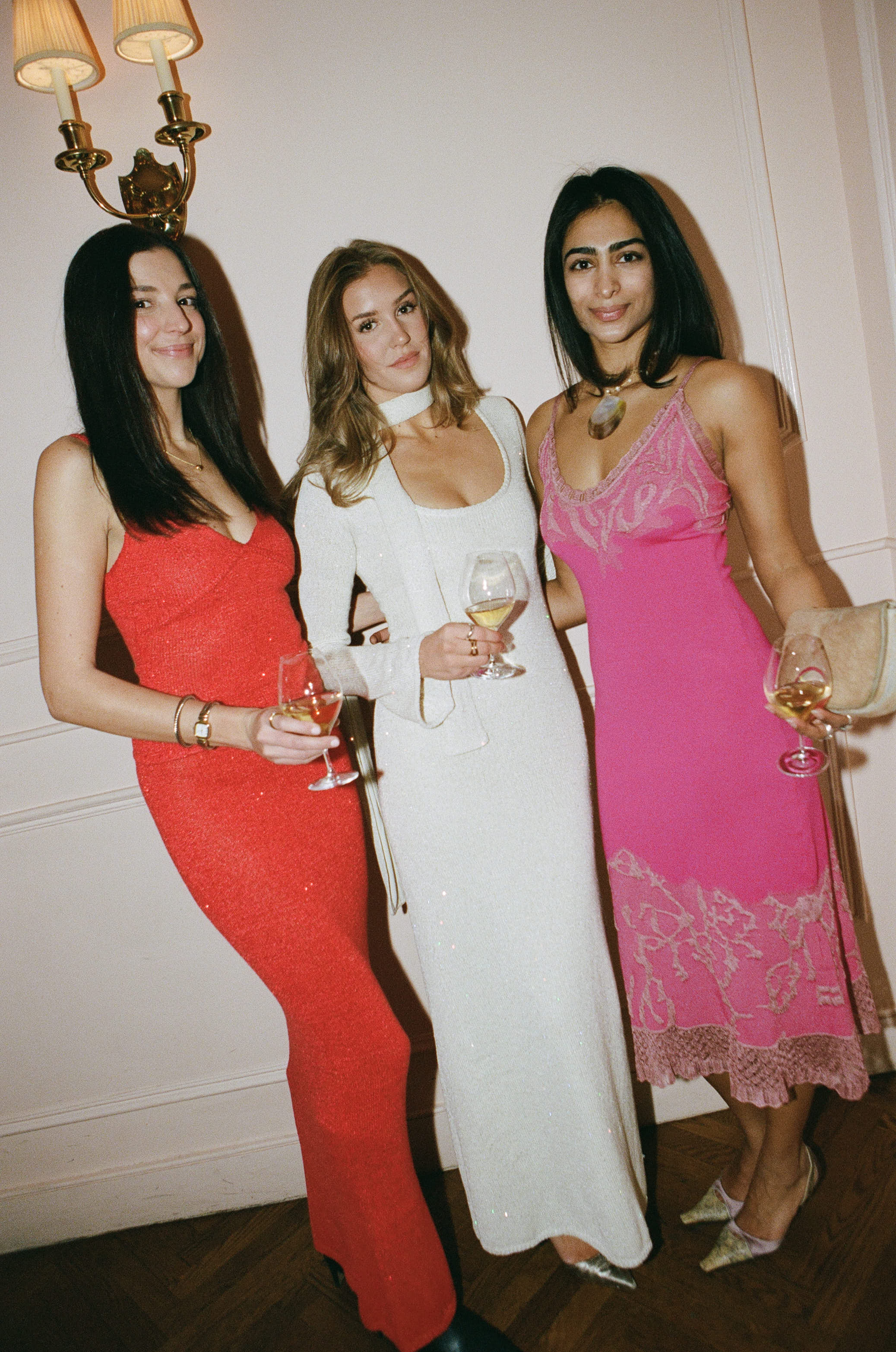 Three women in red, white, and pink dresses holding wine glasses at a party