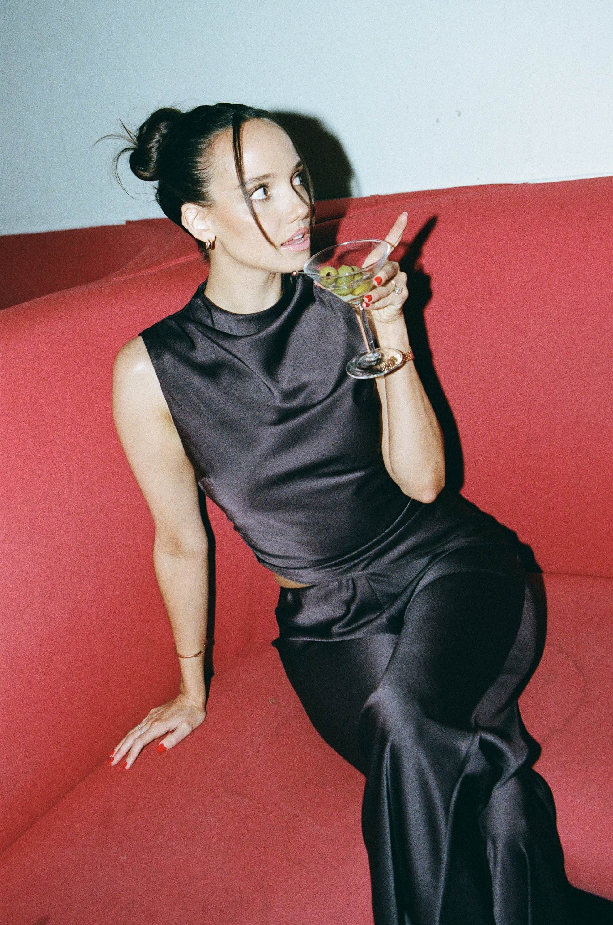 Brunette woman in a black satin set holding an olive martini on a red couch