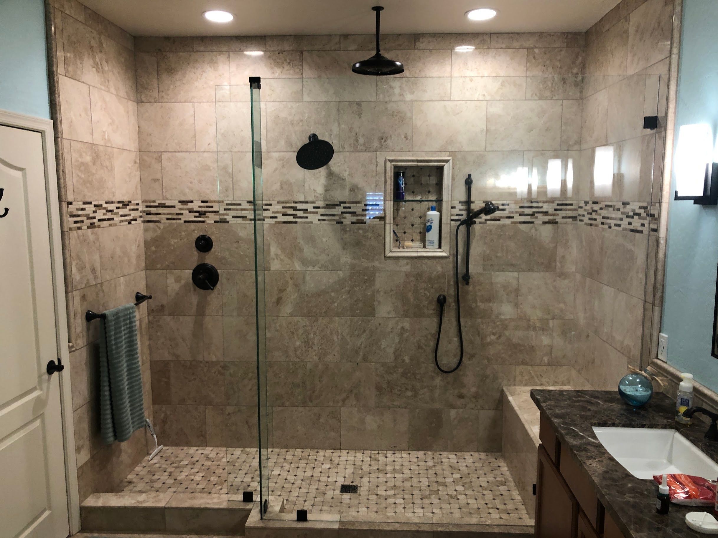Walk-in shower with large-format travertine tiles, decorative mosaic accent band, frameless glass doors, black rainfall fixtures