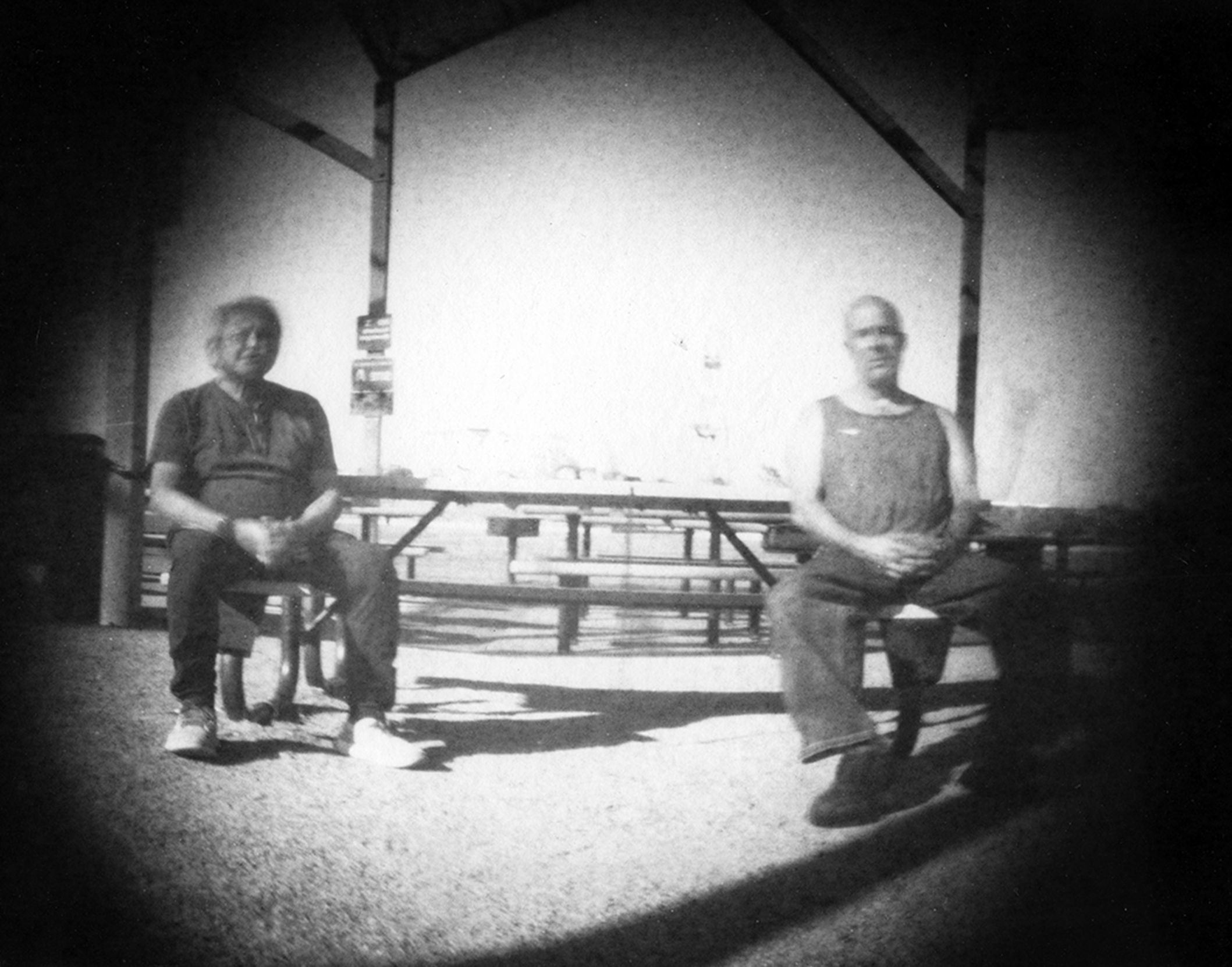Two men seated on chairs in covered pavilion backlit by bright sky with soft focus vignetting and contemplative mood