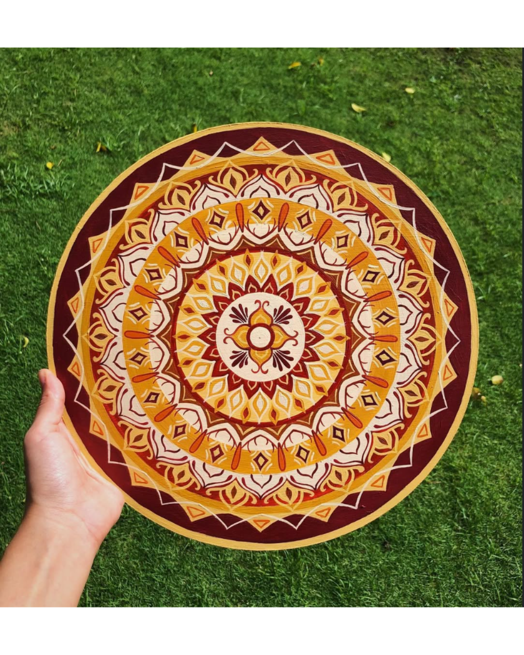 Mandala circular em tons de dourado, laranja e vinho segurada por mão sobre grama verde