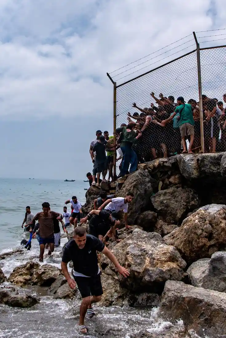 Migrants climbing over a chain-link fence on the rocky Ceuta coastline