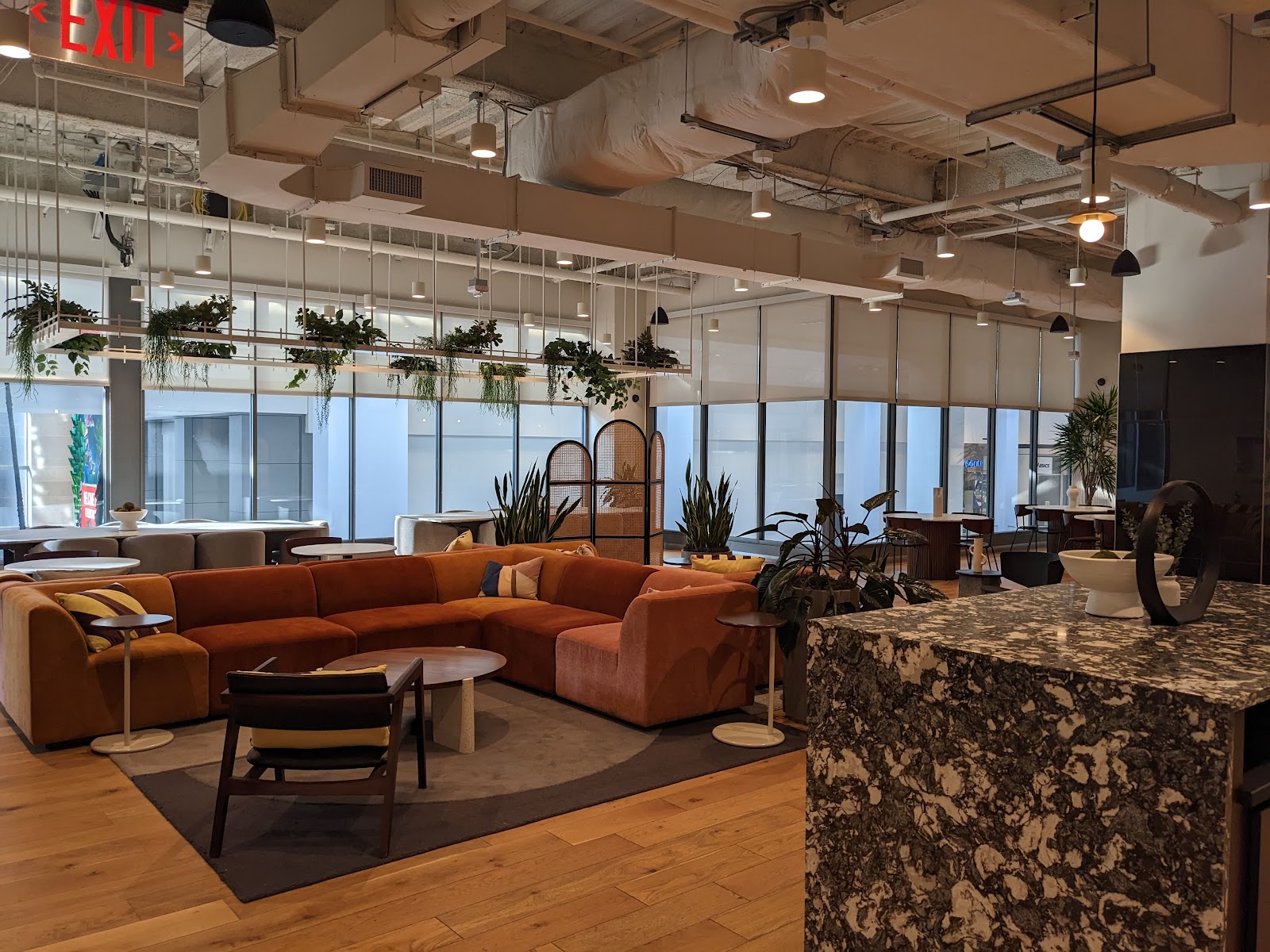 Industrious coworking lounge with burnt orange velvet sectional sofa, granite countertop bar, hanging plants, large floor-to-ceiling windows, modern furniture, and warm earth-tone color palette in upscale hospitality-driven workspace