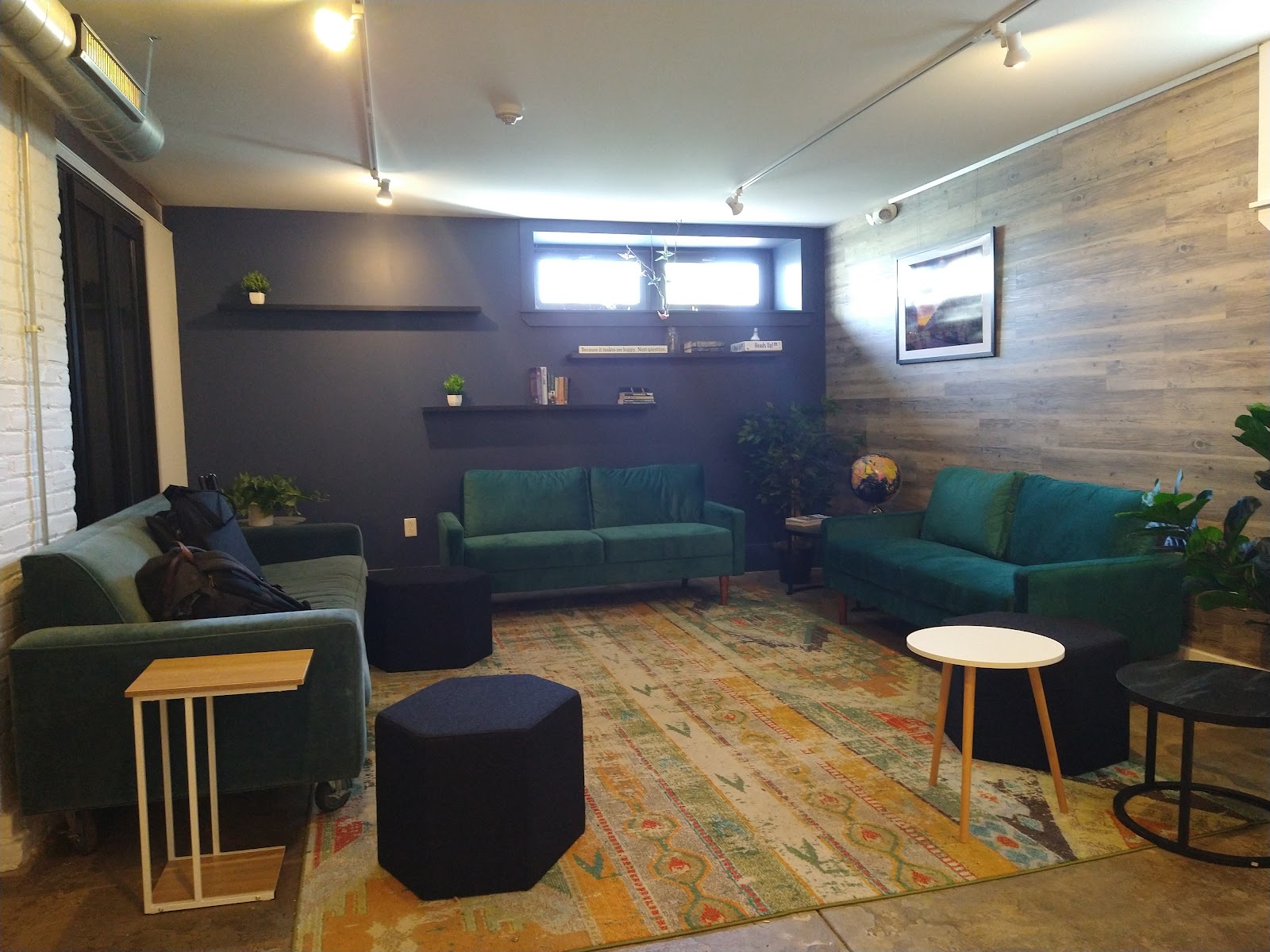 Work Mt. Airy cozy lounge with teal emerald green velvet sofas, dark accent wall with floating shelves, reclaimed wood accent wall, colorful patterned area rug, industrial exposed ductwork, and eclectic community-minded atmosphere