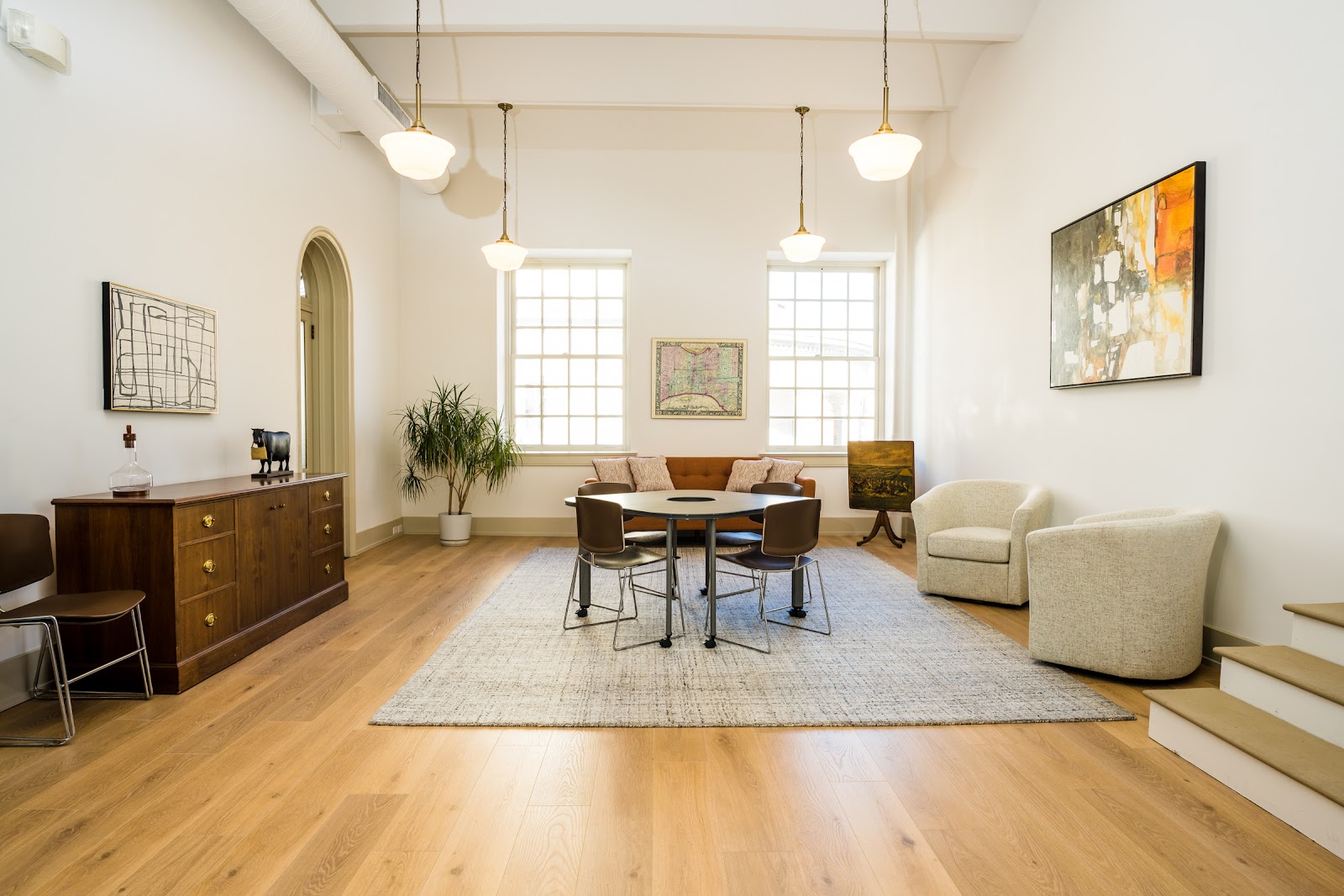 Incutate Workplace Philly elegant high-ceiling space with brass schoolhouse pendant lights, hardwood floors, arched doorway, vintage-inspired furniture, antique credenza, modern abstract art on walls, and sophisticated gallery-like atmosphere in historic Old City building