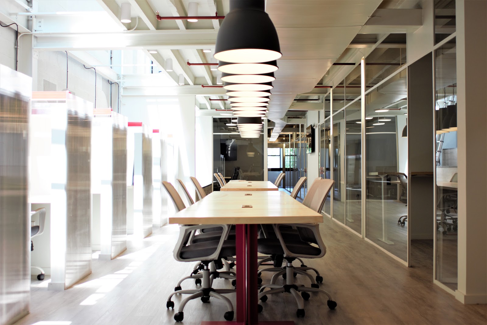 Billy Penn Studios Coworking industrial modern workspace with exposed white ceiling and ductwork, modern tiered pendant lights, long wooden communal table with office chairs, glass-walled private offices, bright natural light, and professional collaborative environment in converted Fishtown building
