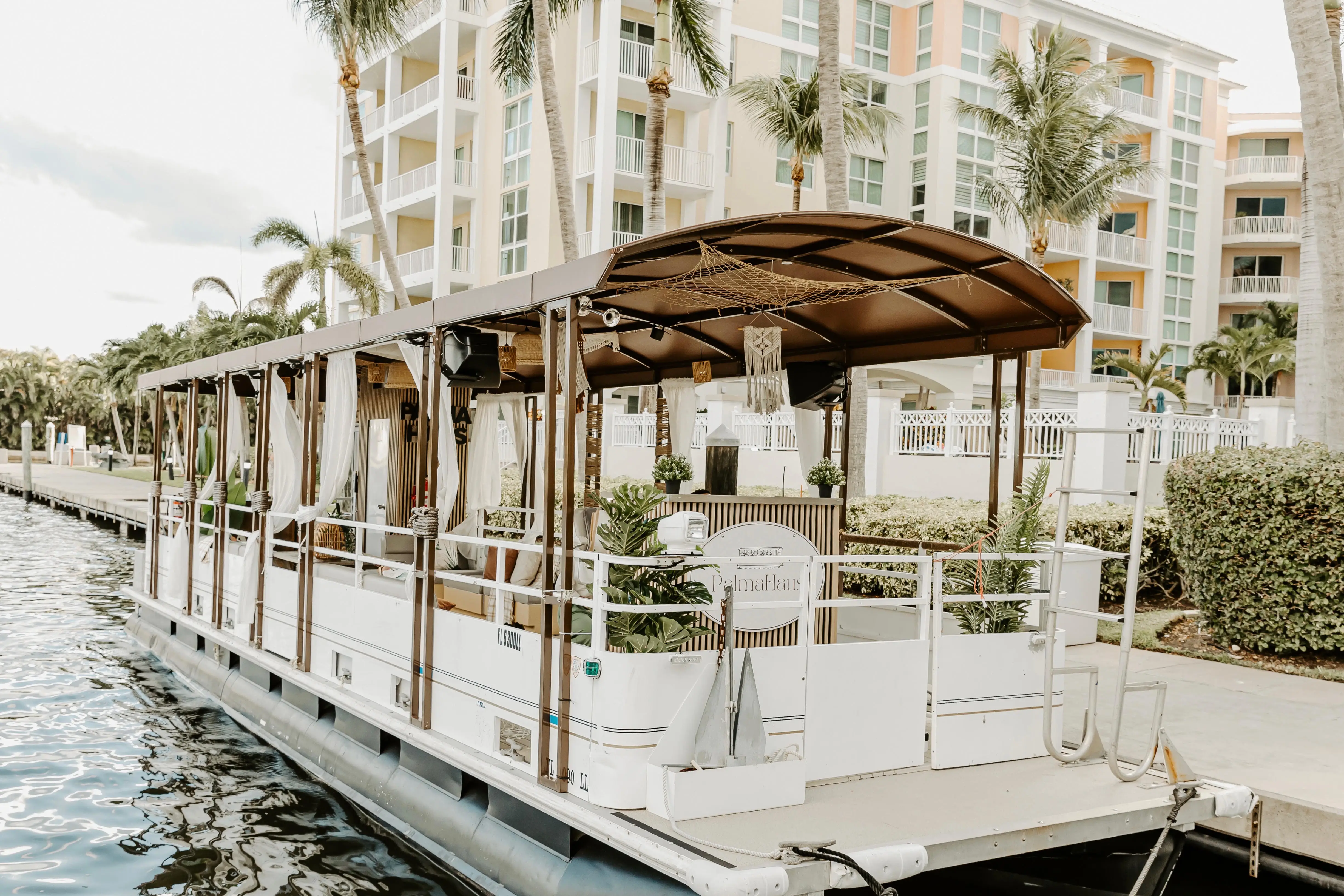 The Elite Sport Yachts 45ft Pontoon 'PalmaHaus' moored along a West Palm Beach waterway canal, showing its full white hull, sweeping brown curved roof canopy, open-air sides with white sheer curtains, and lush tropical plant decor