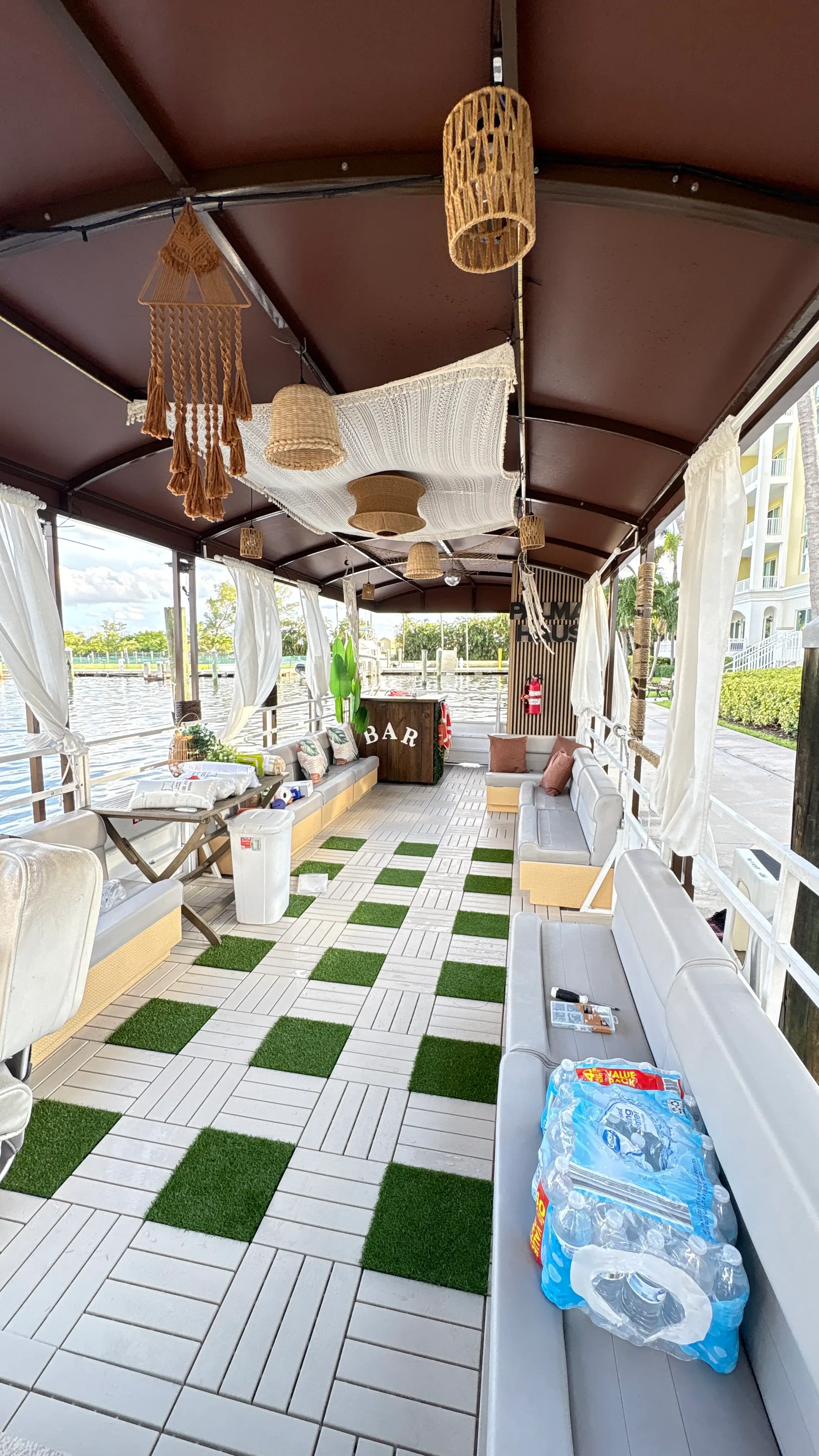 Wide interior view of the Elite Sport Yachts 45ft Pontoon looking down the full length of the covered deck, showing rattan and macramé hanging décor, white sheer curtains, gray bench seating along both sides, and the wooden BAR station